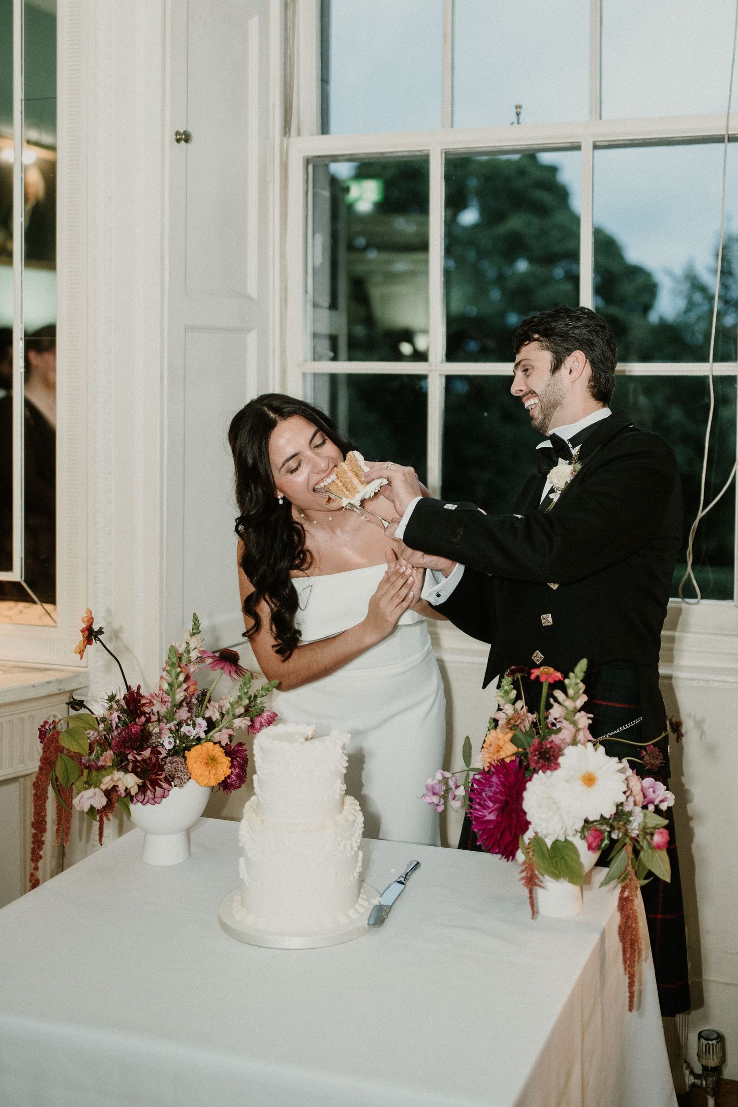 Wedding cake cutting moment in a bright sunlit room with floral arrangements.
