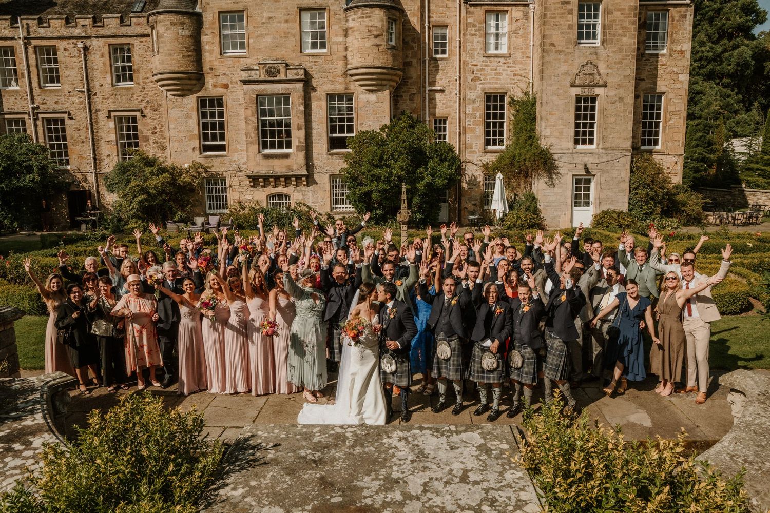 Large group wedding photo in front of historic stone mansion with formal gardens.