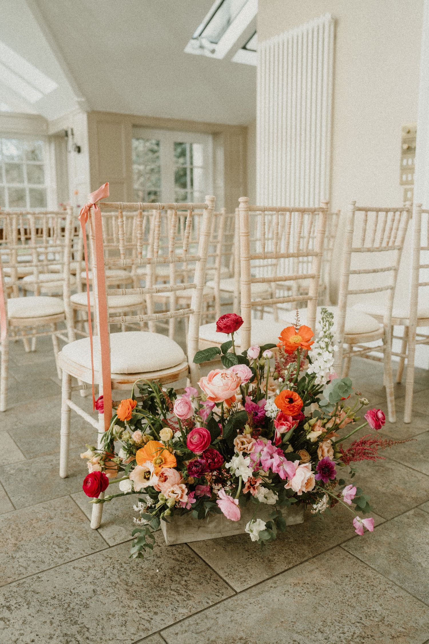 Elegant floral arrangement with pink and orange blooms decorates a wedding ceremony space with light wooden chiavari chairs.