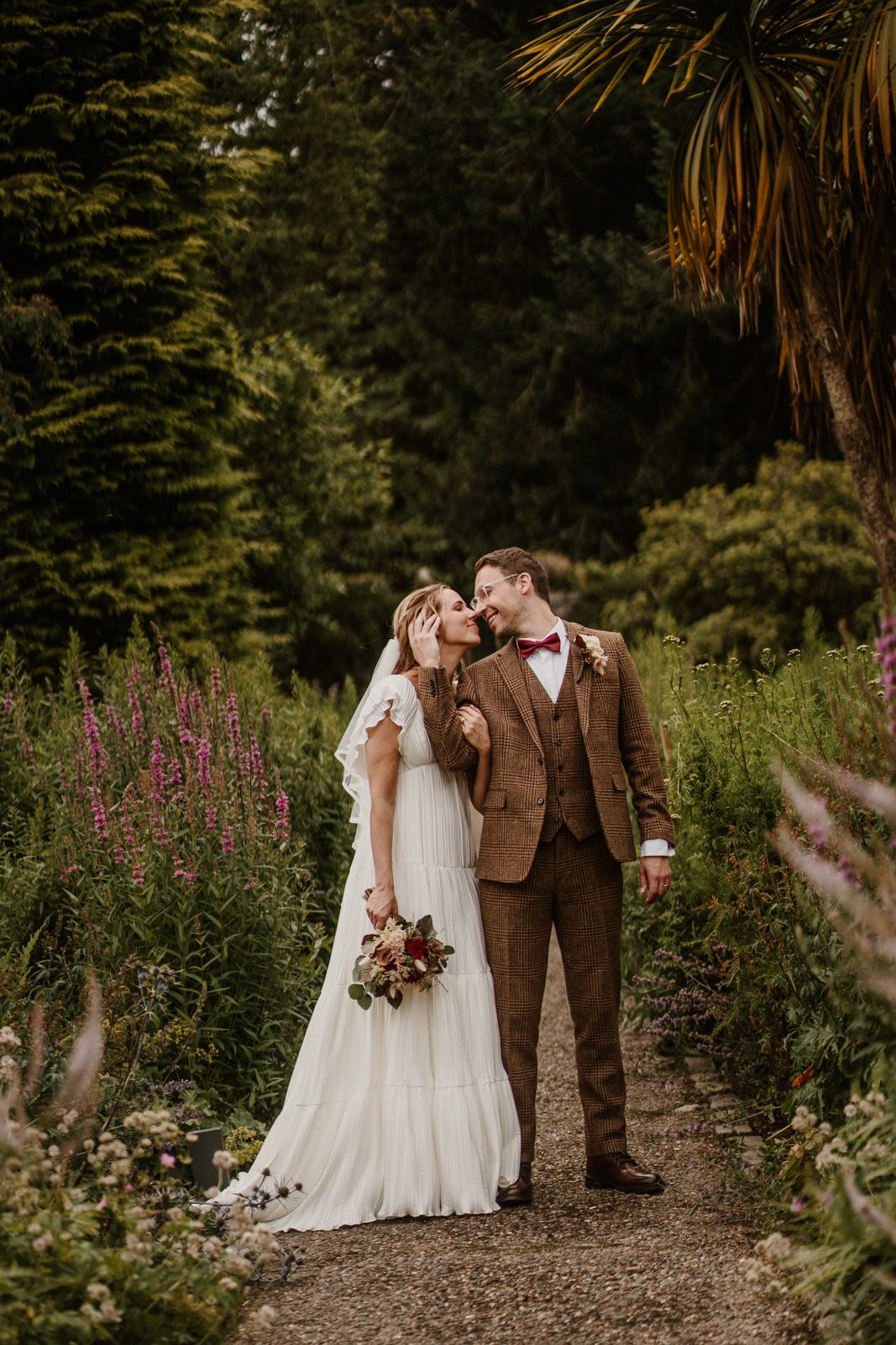 Romantic garden portrait with couple in brown suit and white dress among wildflowers.
