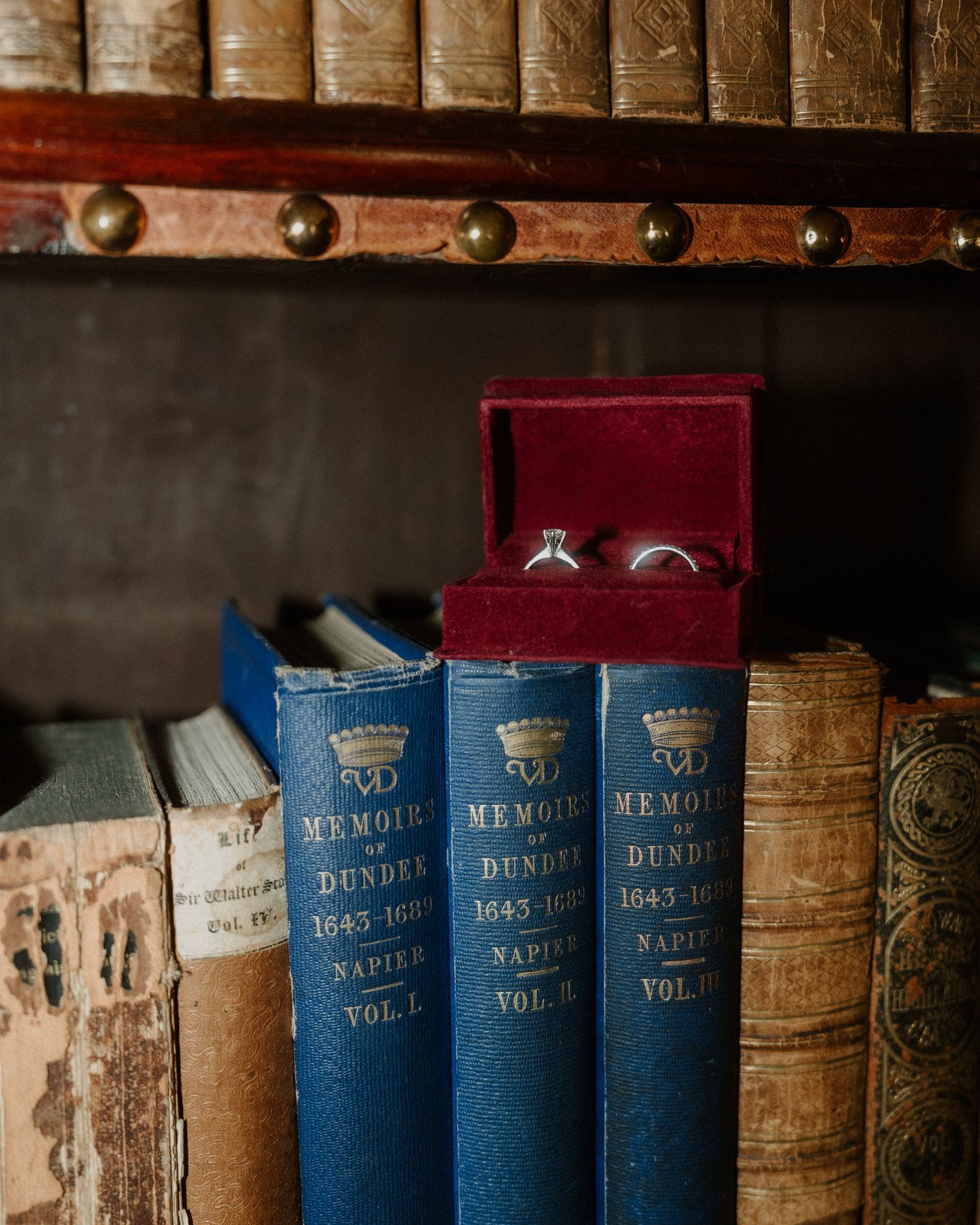 Wedding rings in a red velvet box displayed on vintage blue Memoirs of Dundee books on a wooden shelf.
