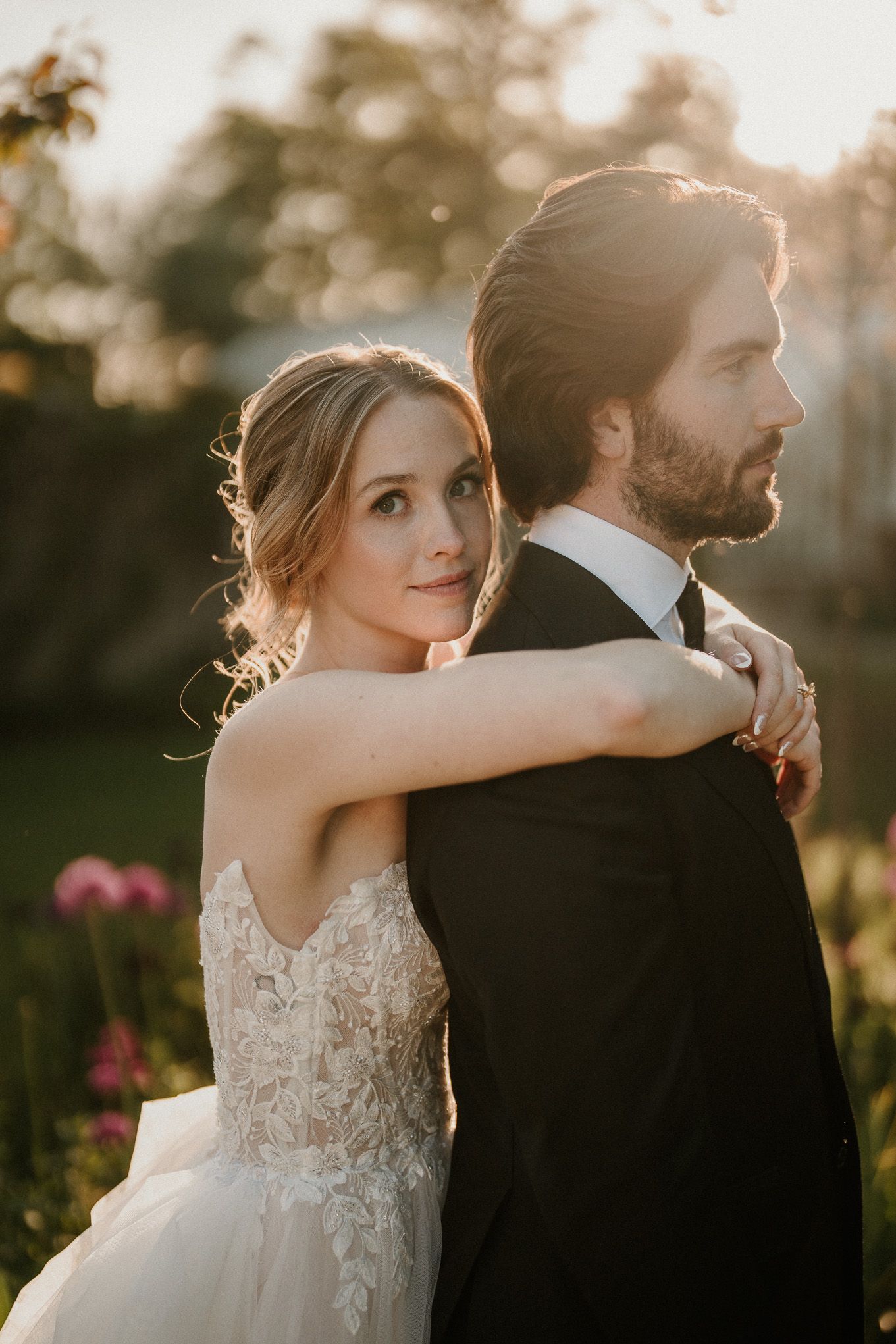 A bride in lace gown embraces a bearded groom in black tuxedo during golden hour in a garden setting.
