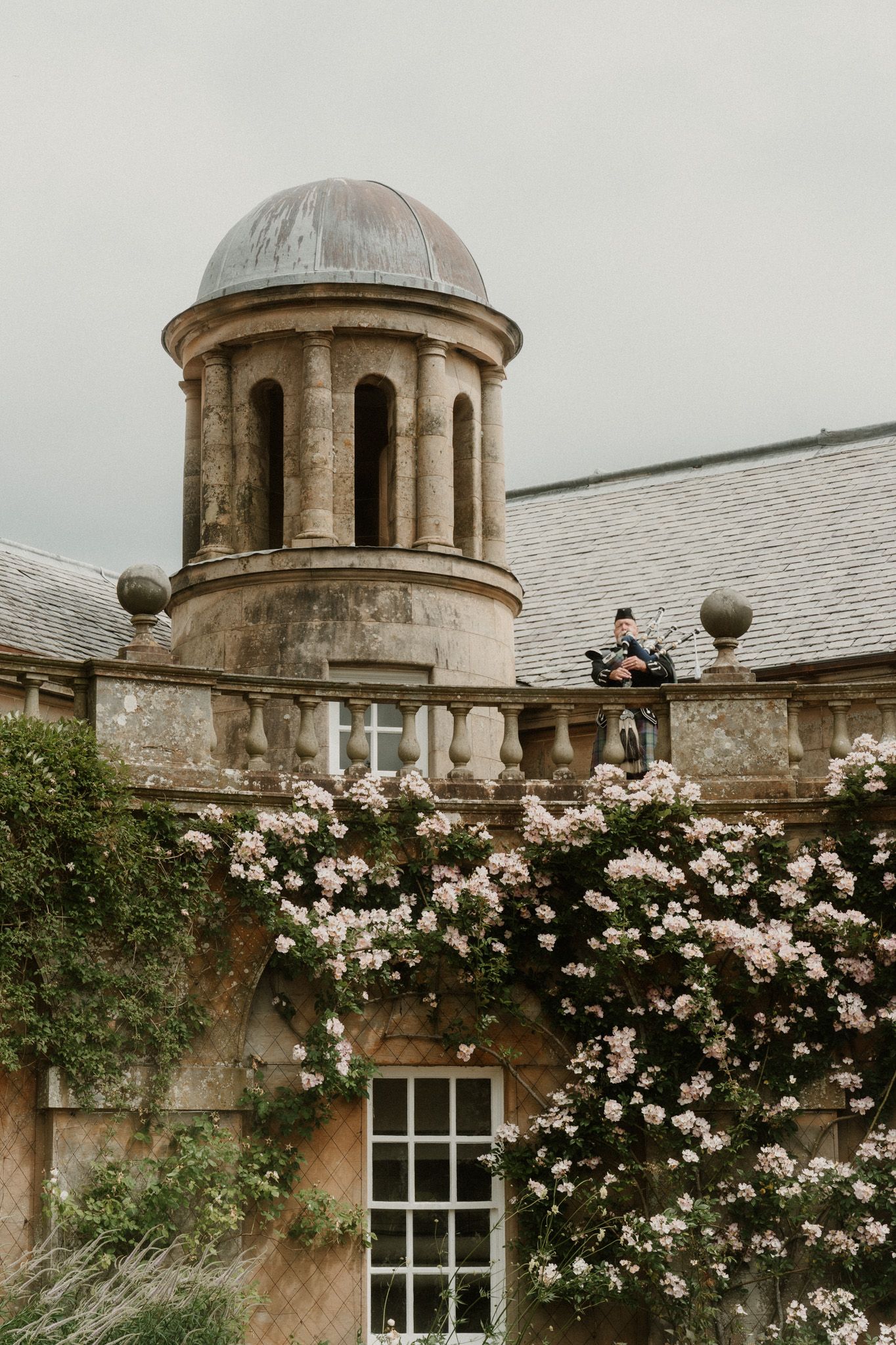 Bagpiper plays atop a stone balcony adorned with climbing pink roses on a historic estate with domed tower.