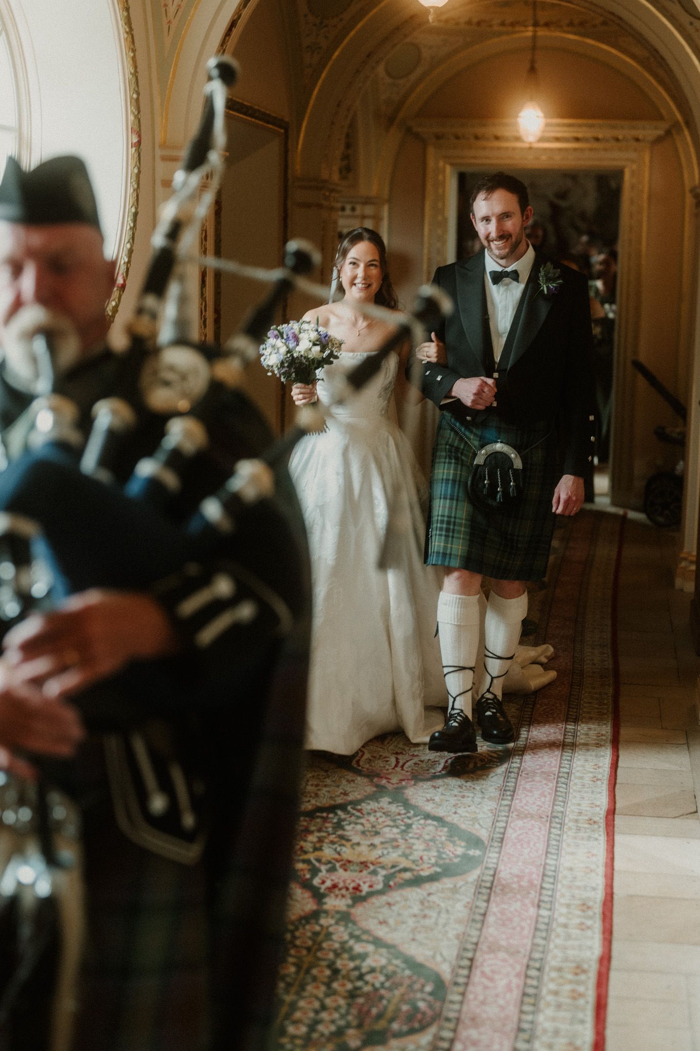 Scottish wedding couple walks down hallway led by bagpiper, groom in tartan kilt, bride holding bouquet.