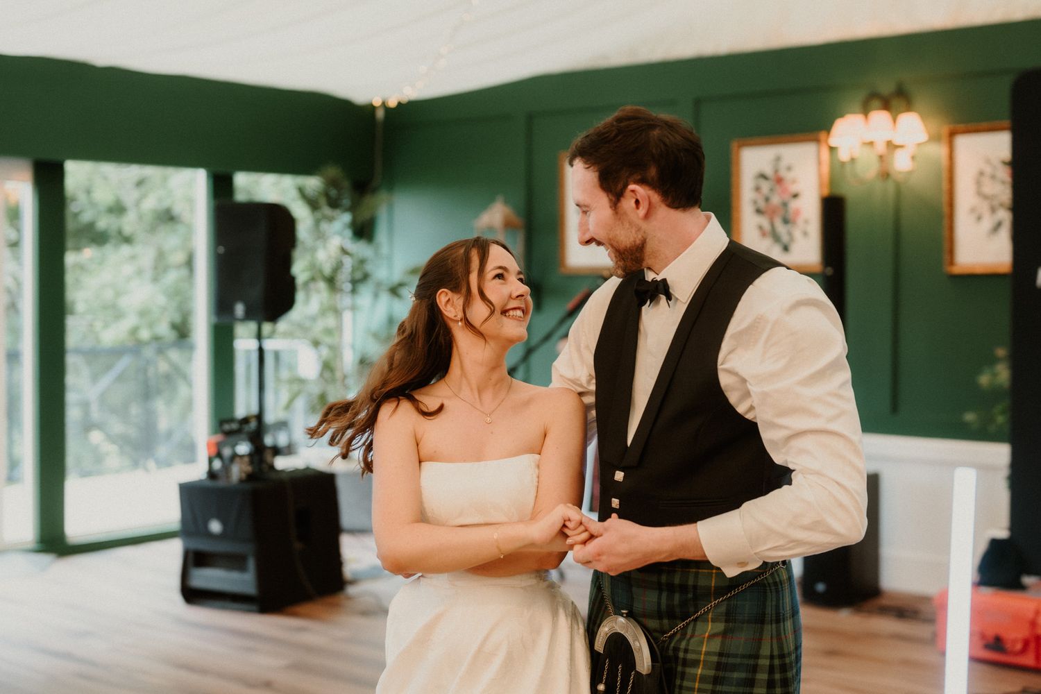 Bride and groom share first dance in green-walled marquee, groom wearing traditional tartan kilt.