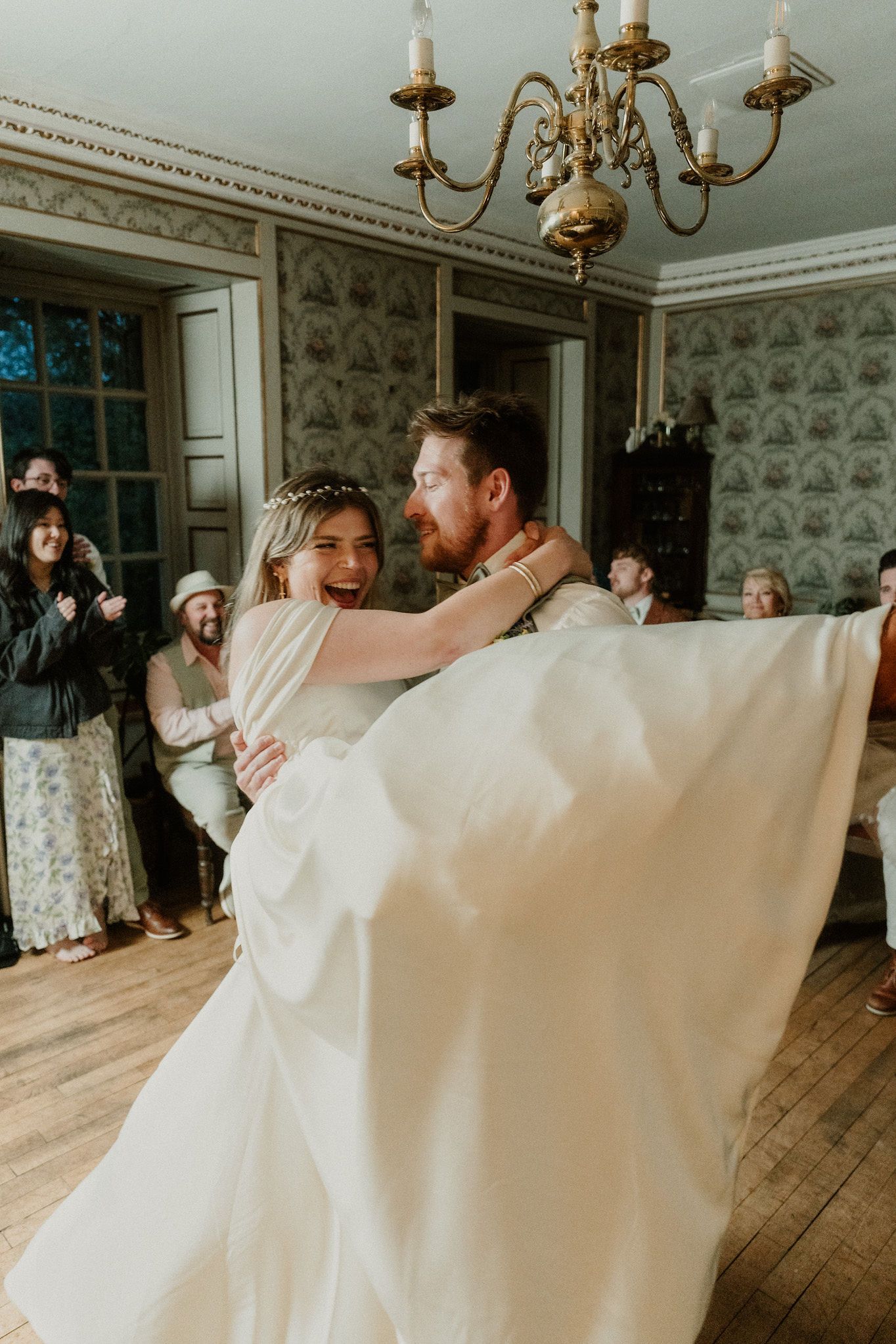 A joyful bride and groom share their first dance in an elegant vintage room while wedding guests watch and celebrate.