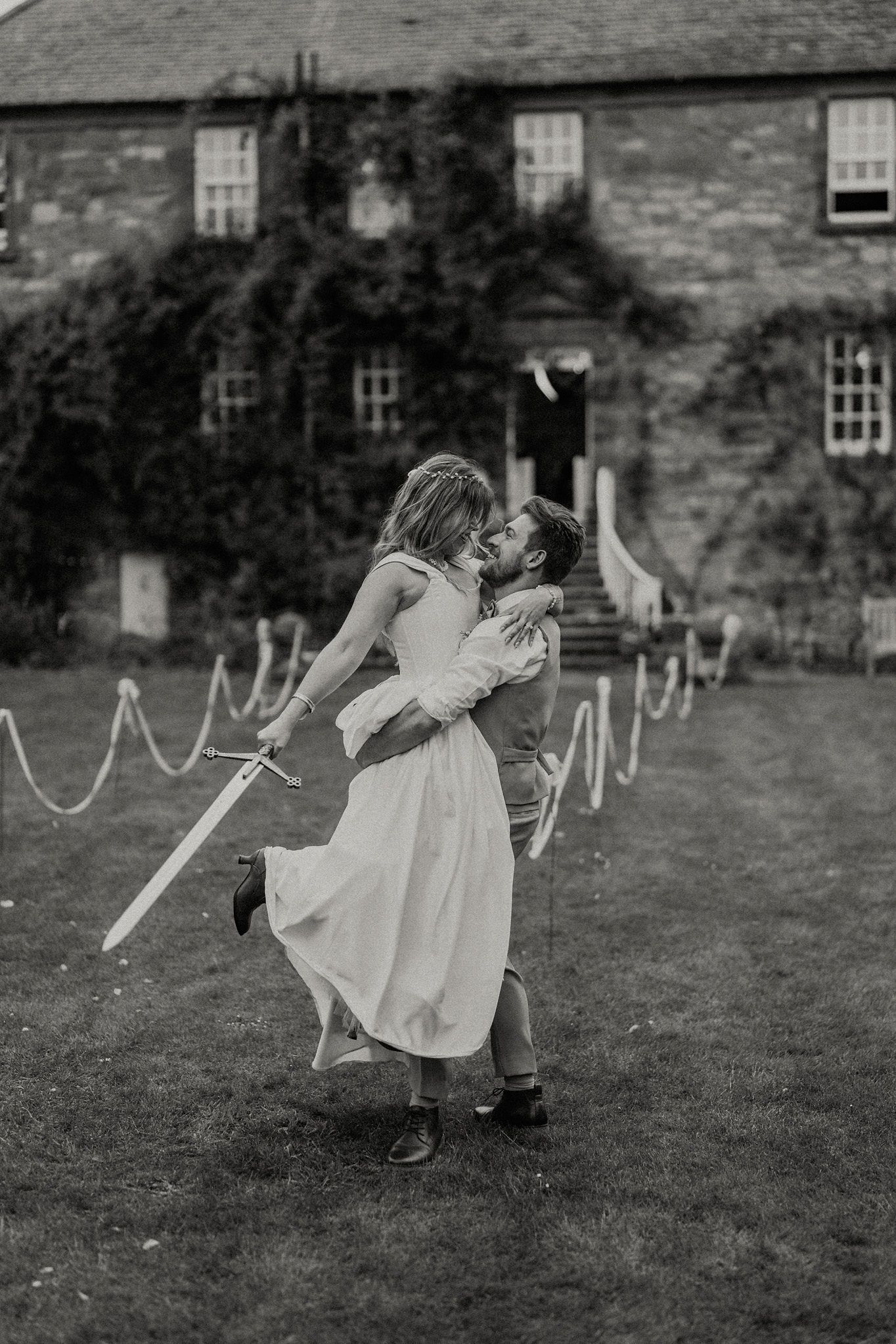 Black and white photo of a couple kissing while the groom lifts the bride holding a sword outside a stone manor.