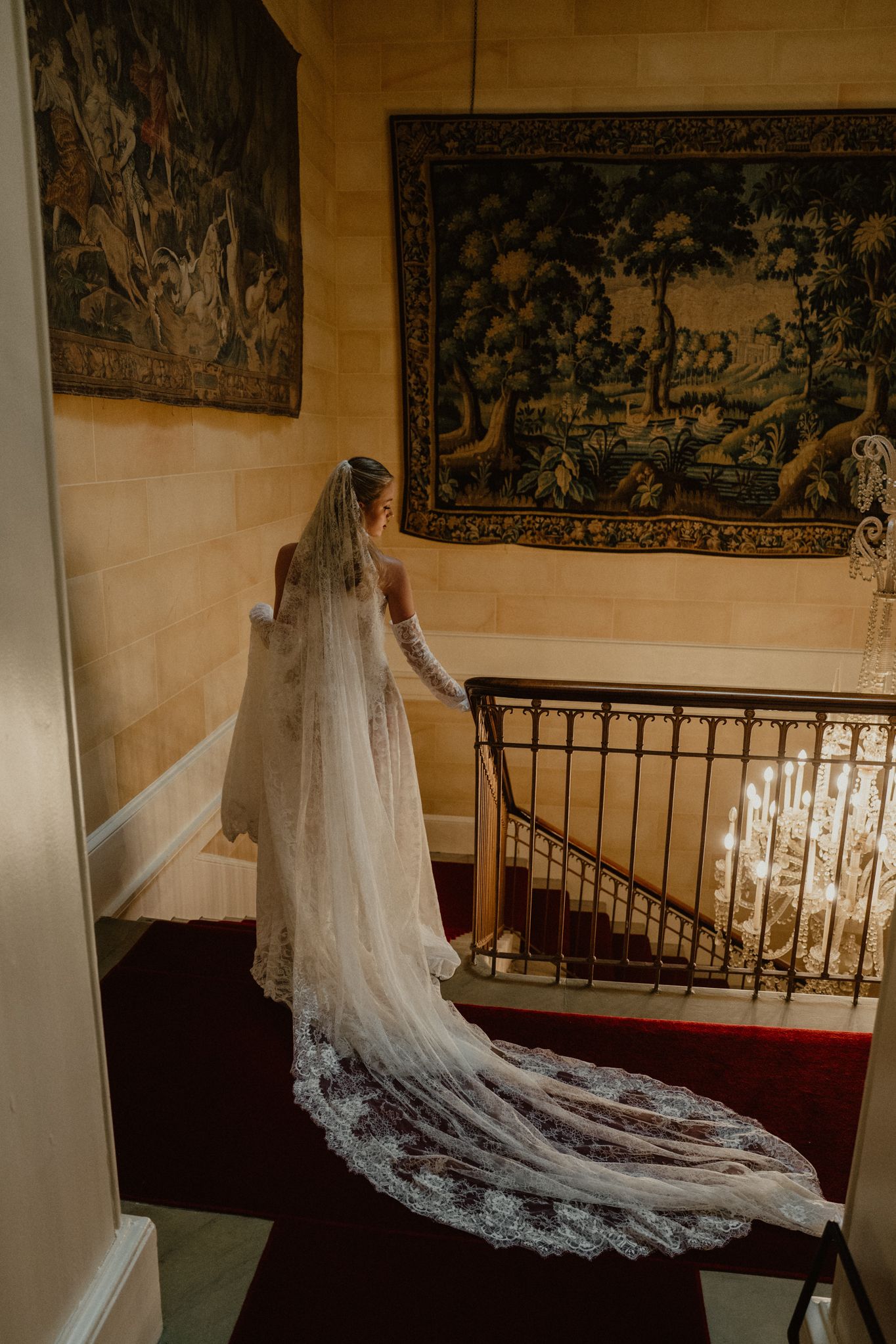 A flowing white wedding dress with a long lace train photographed on a staircase with tapestries in the background.