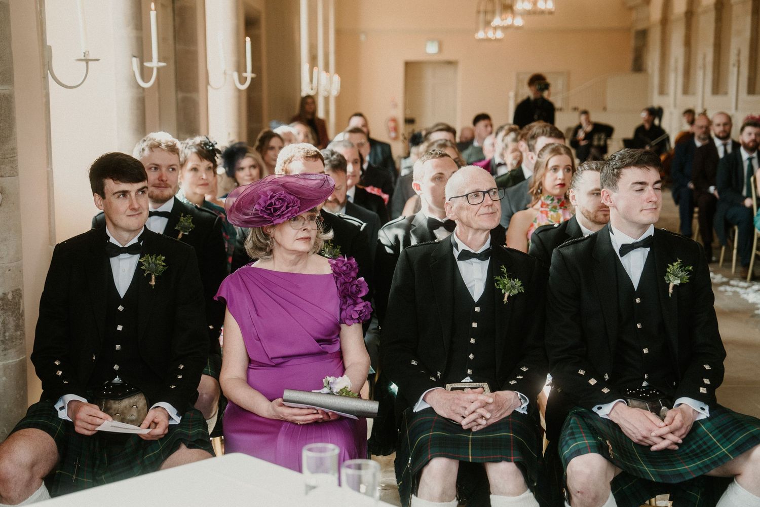 Wedding guests in formal attire and kilts seated in rows during an indoor ceremony.