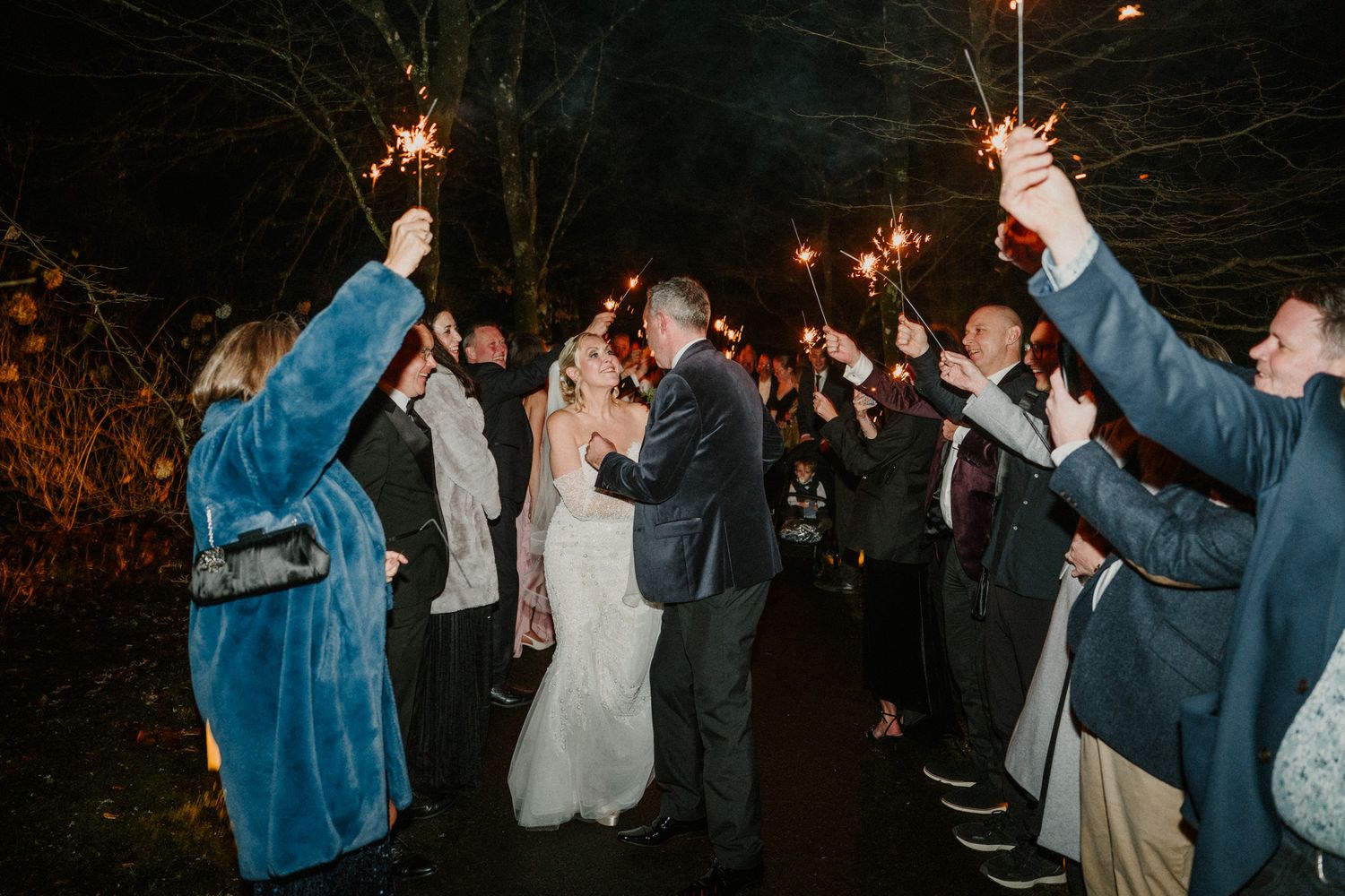 Wedding party dances and celebrates with sparklers creating a festive outdoor atmosphere.