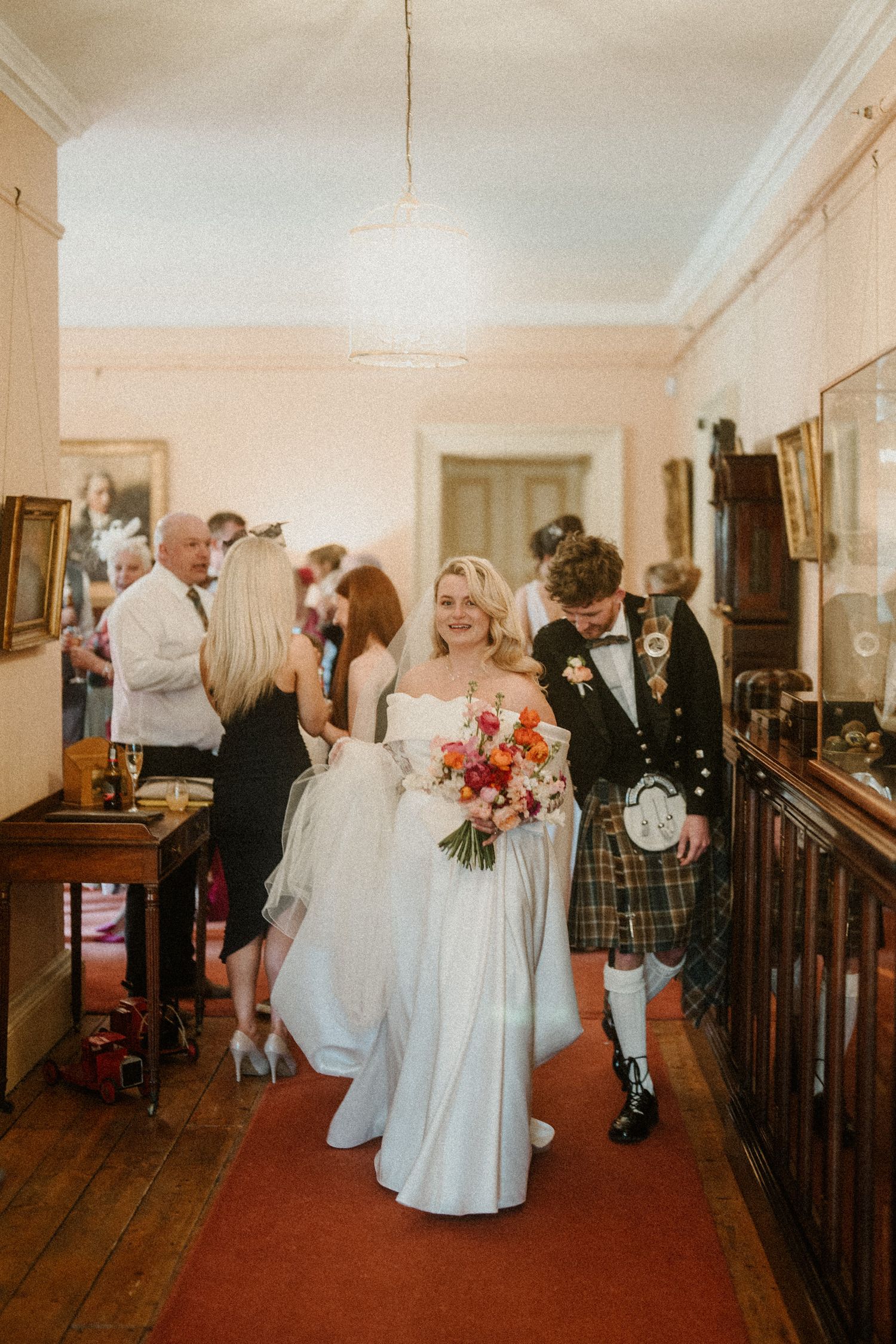Wedding party members walk through an elegant indoor venue with wooden floors and traditional decor.