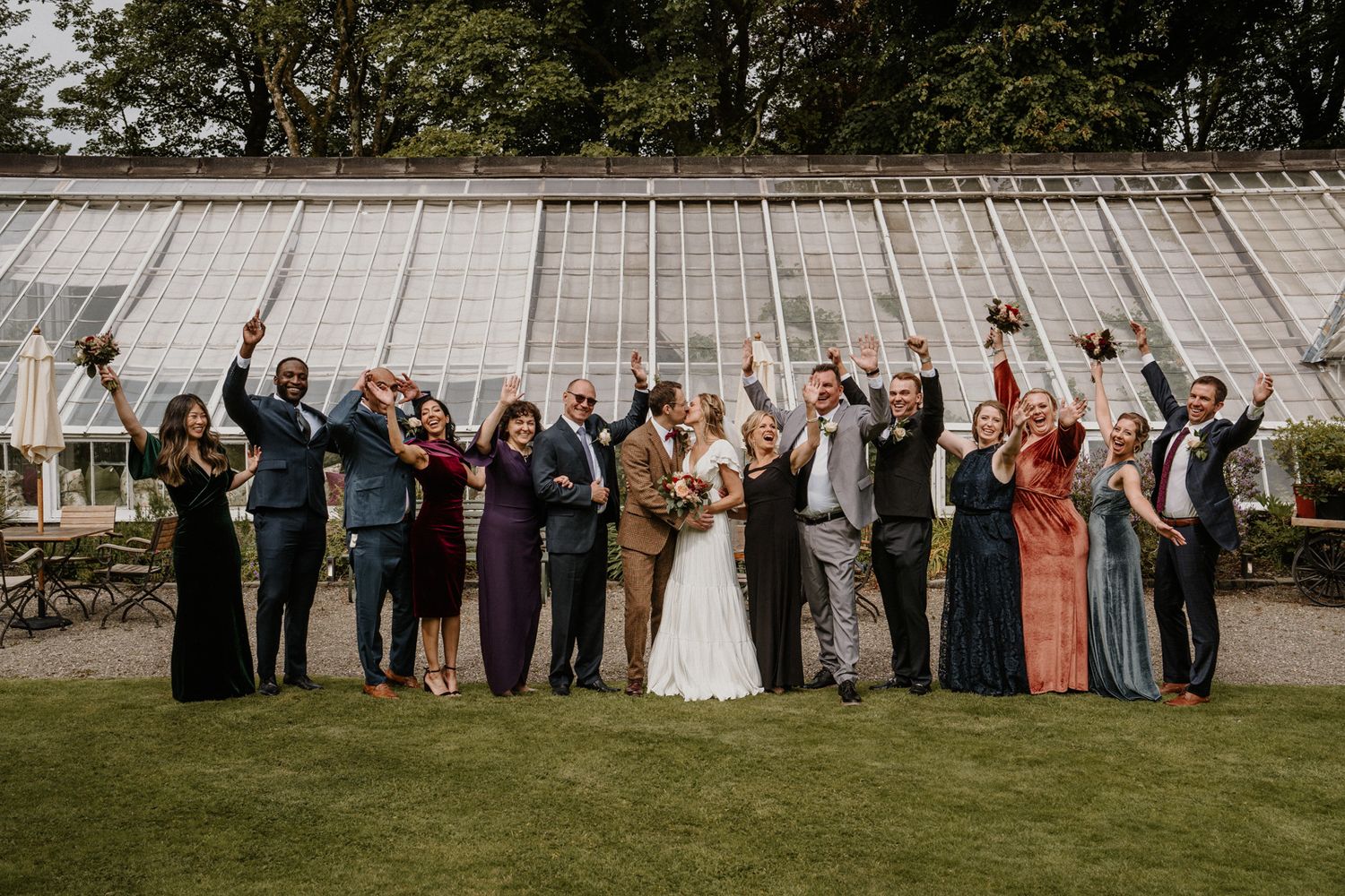Wedding party and guests celebrating with raised arms in front of greenhouse.