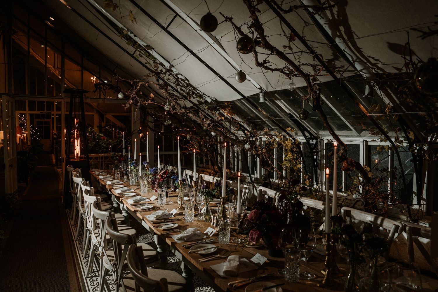 Dramatic evening reception table setting with candles and place settings in a glass-enclosed venue.