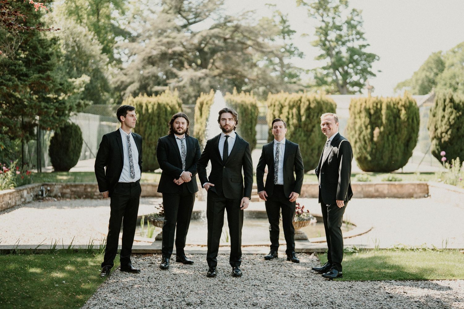 Wedding party in black suits standing on gravel path surrounded by lush garden scenery.