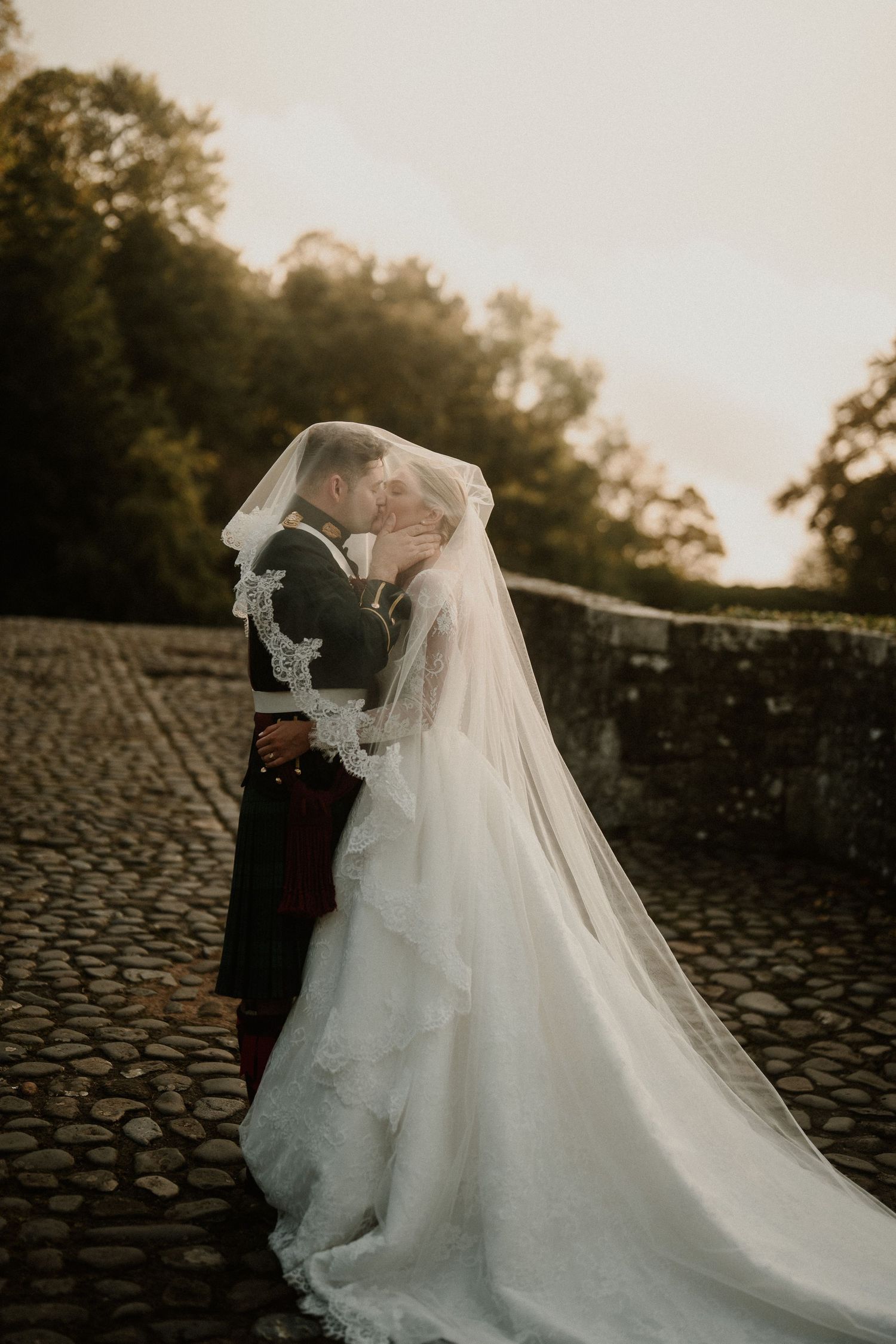 Bride and groom share romantic kiss under lace veil on cobblestone path at golden hour outdoor wedding.