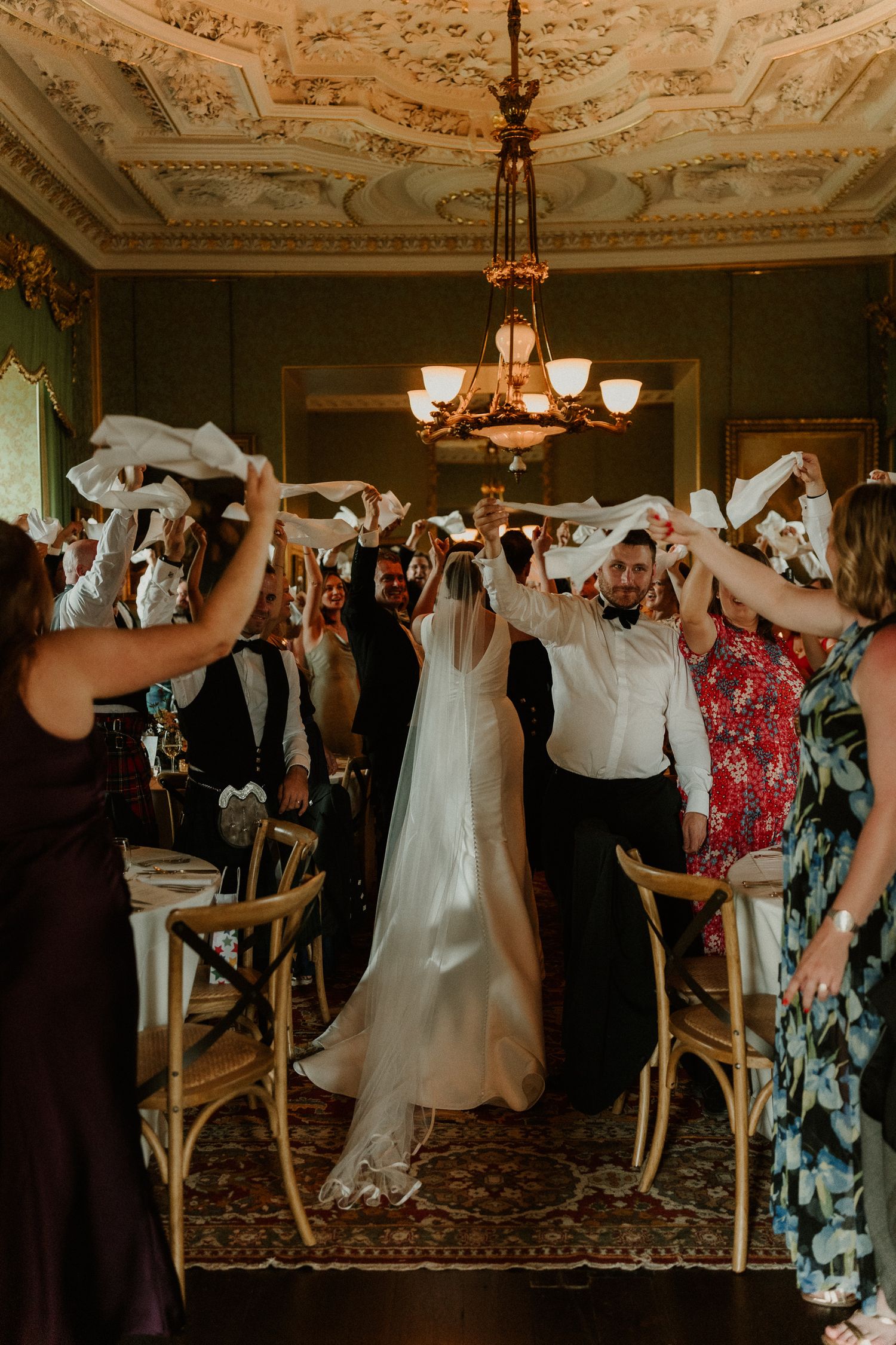 Bride and groom walk through napkin wave tunnel at elegant wedding reception with ornate ceiling.