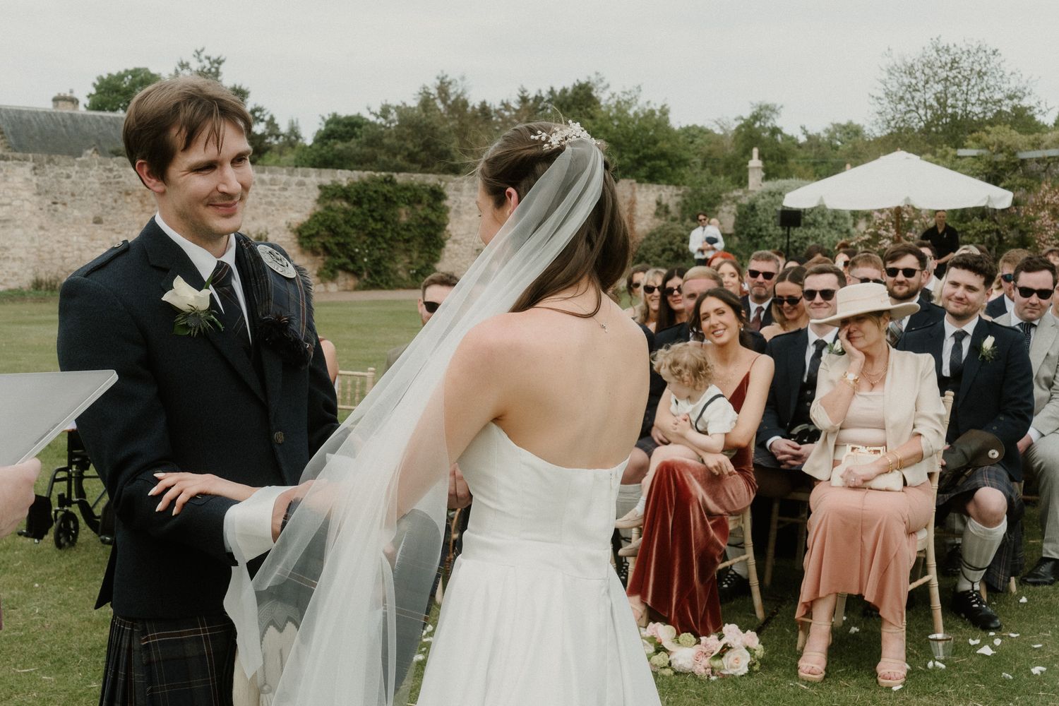 Groom in kilt smiles at bride in white gown and veil during outdoor wedding ceremony with seated guests.