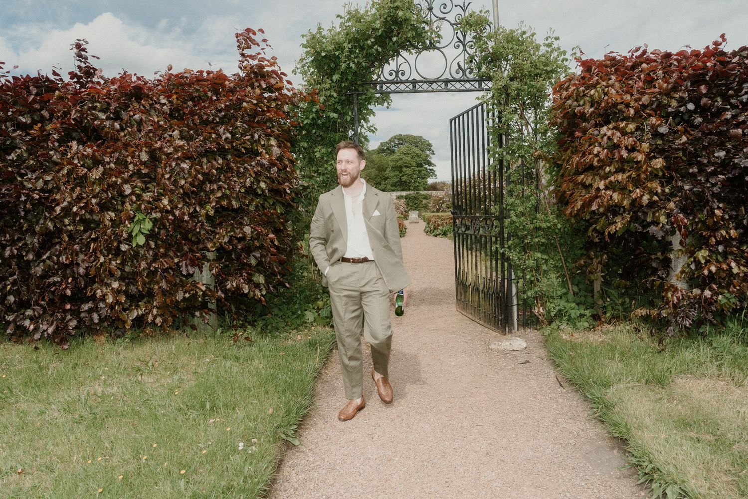 Man in sage green suit walking through ornate iron garden gate flanked by tall red-leafed hedges on a gravel path.