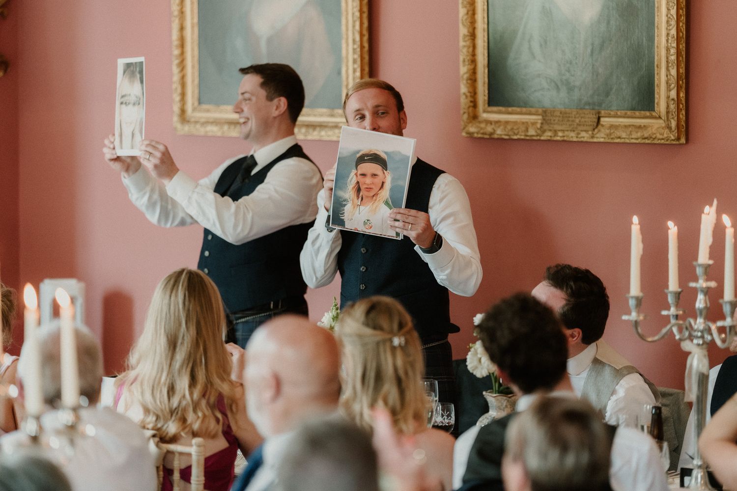 Two groomsmen hold up photos during a wedding reception speech in an elegant pink-walled dining room with candles.
