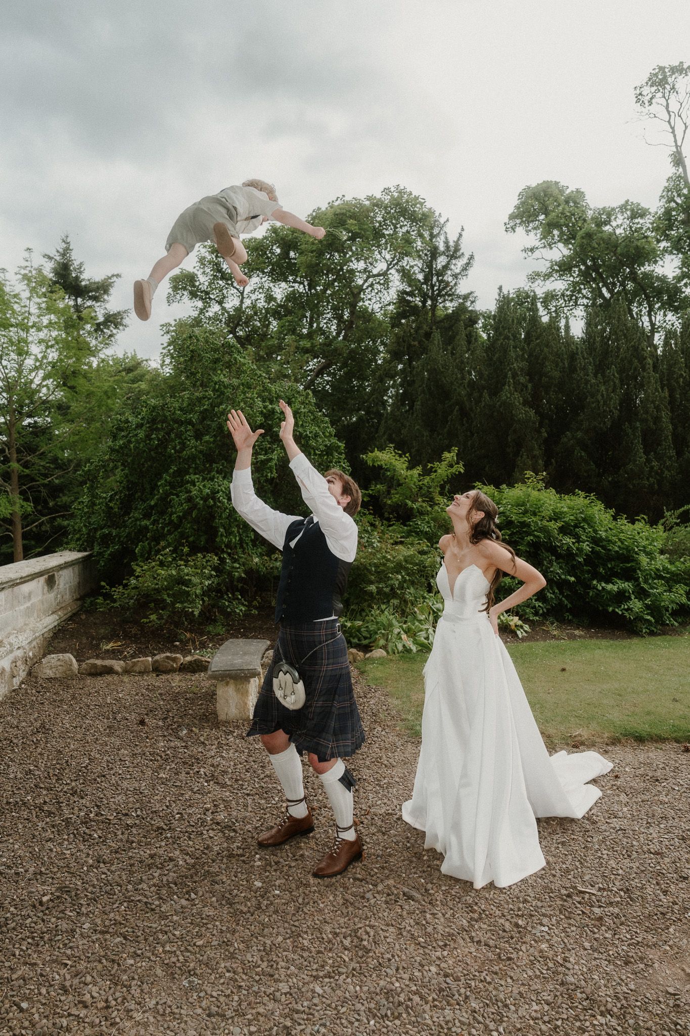 Groom in kilt tosses child in air as bride in white gown laughs at outdoor wedding celebration.