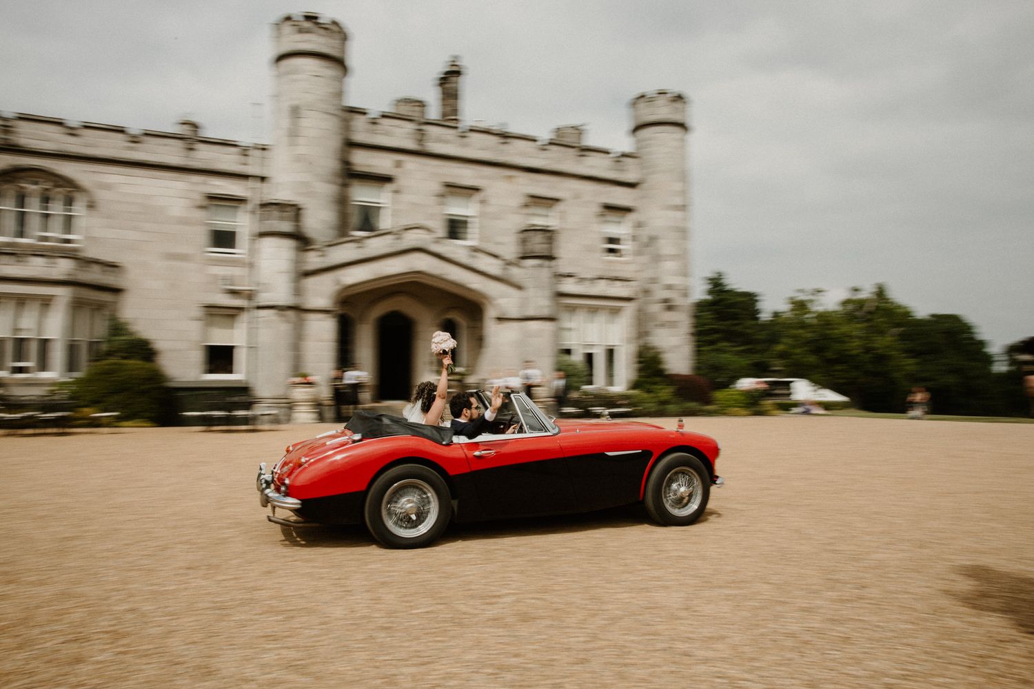 Bride and groom driving a classic red Austin-Healey convertible sports car in front of a grand castle venue.