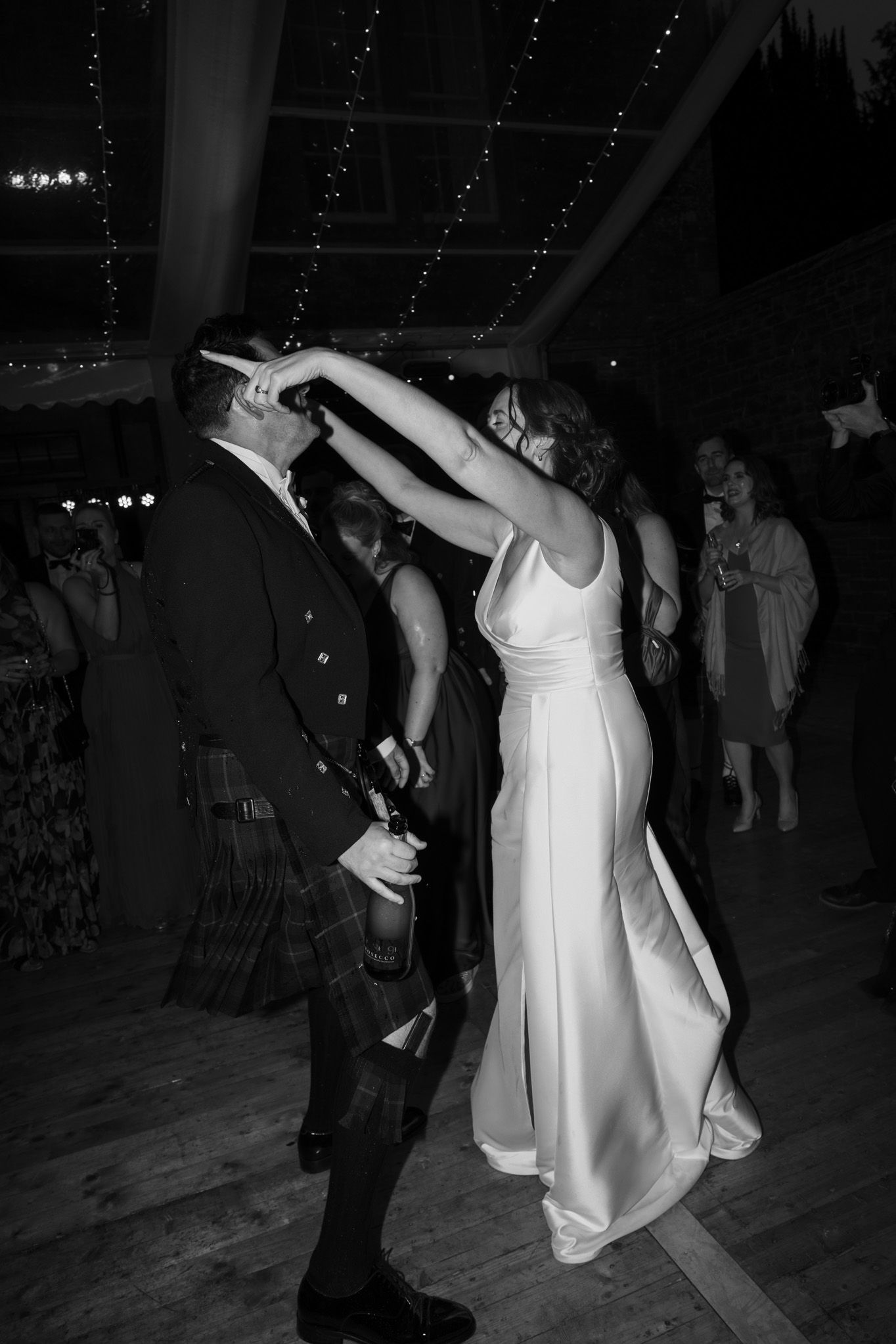 Bride in a satin gown twirls under her partner's raised arm while he wears a kilt, dancing at a candid black and white wedding reception.