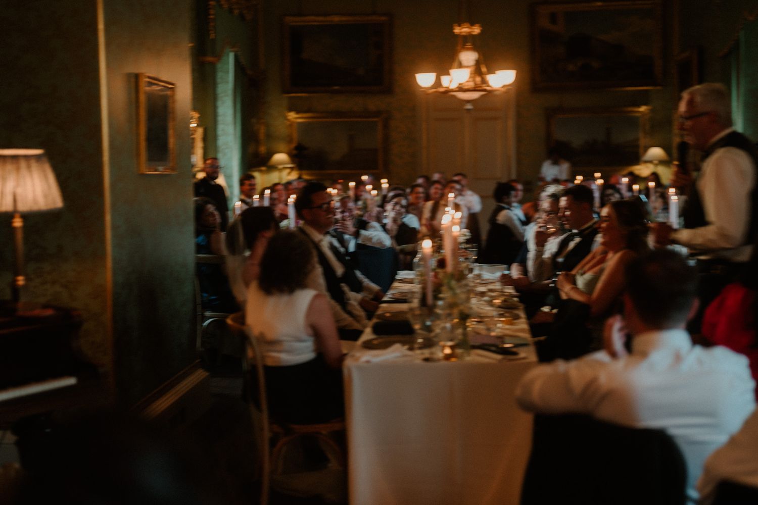 Guests in formal attire dine at a long candlelit table in an elegant, dimly lit room with a chandelier and framed paintings.