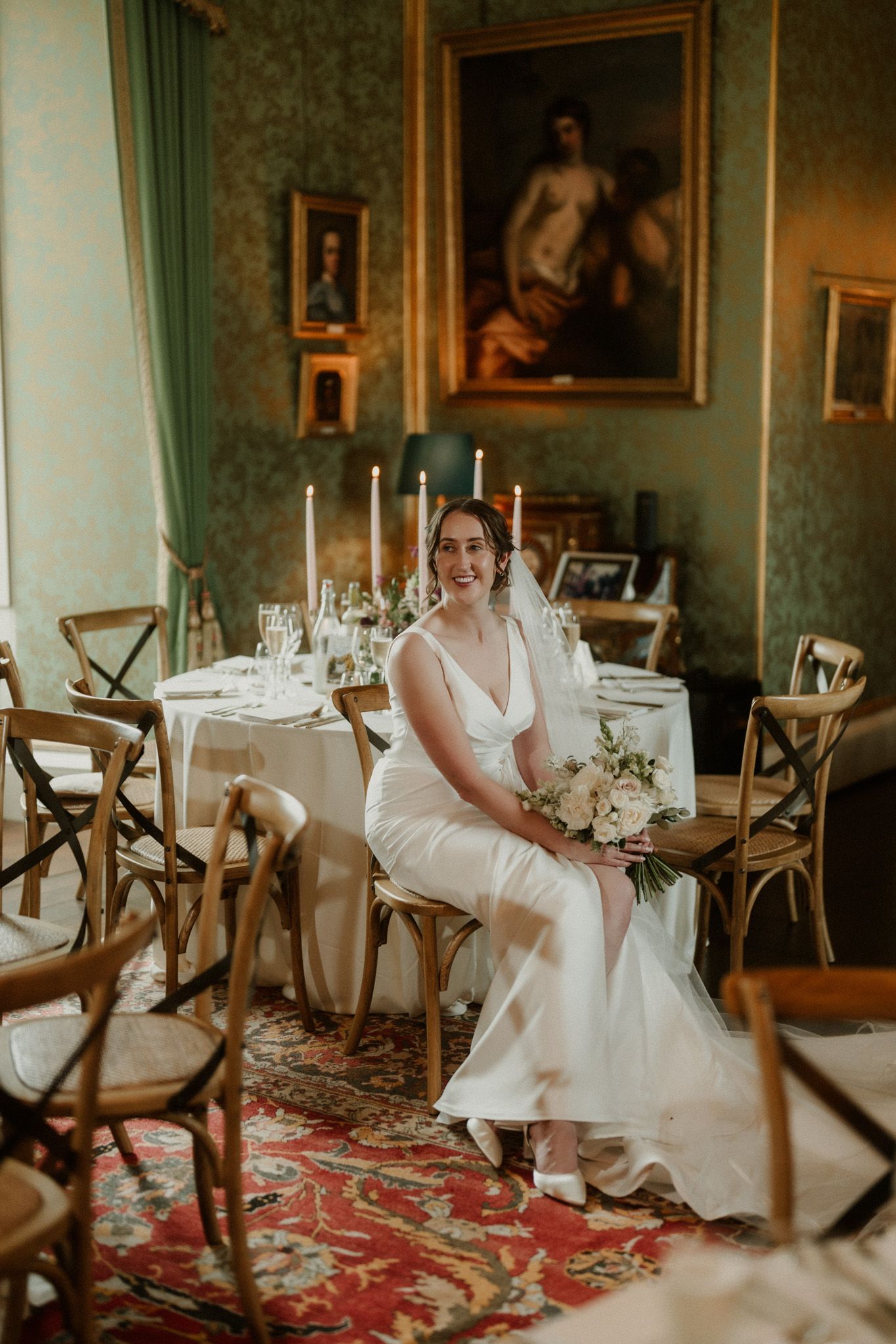 Smiling bride in an elegant white gown sits holding a bouquet in a grand green dining room with candlelit tables and a red patterned rug.