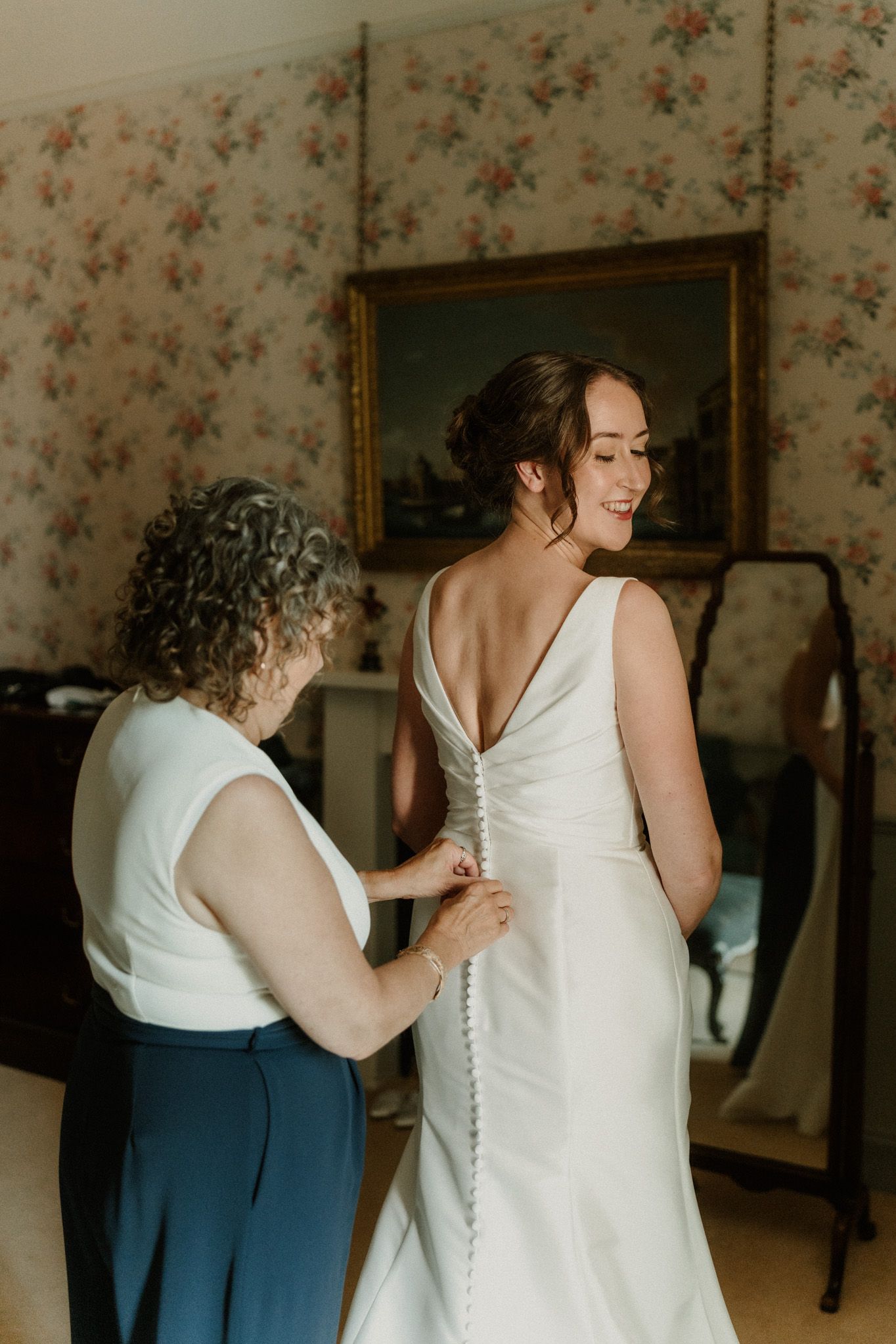 A woman in an elegant white wedding gown smiles as an older woman fastens the row of fabric-covered buttons down the back of the dress.