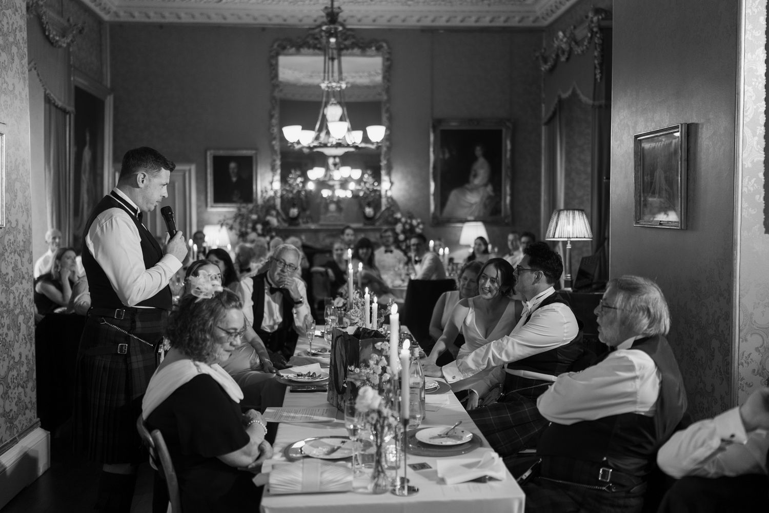 Man in a vest stands giving a speech at a long candlelit dining table as guests in an ornate room with chandeliers look on.