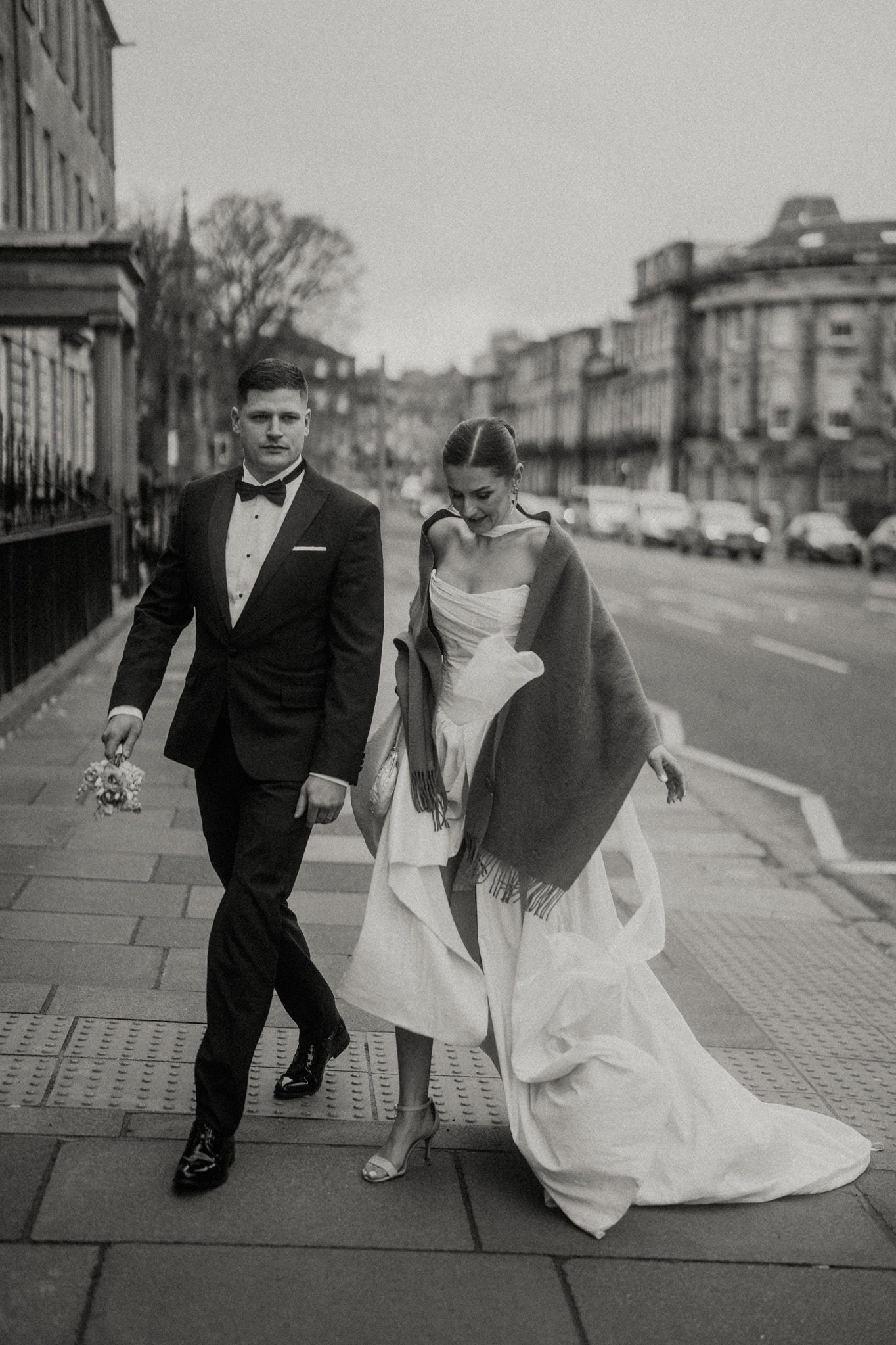 A romantic black and white photo series of a couple walking down a city sidewalk in wedding attire.