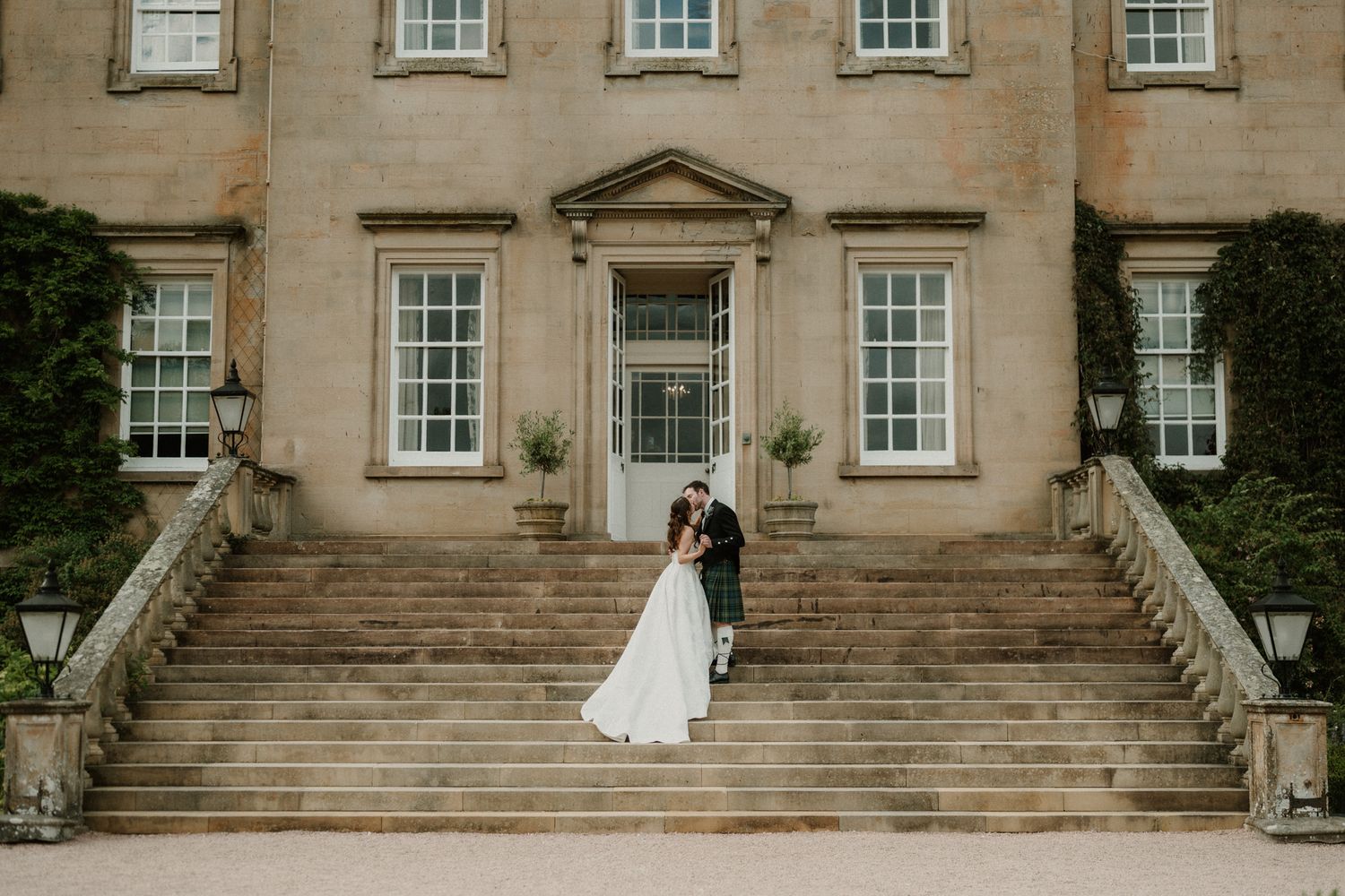 Bride and groom embrace on grand stone steps of elegant Georgian manor house wedding venue.