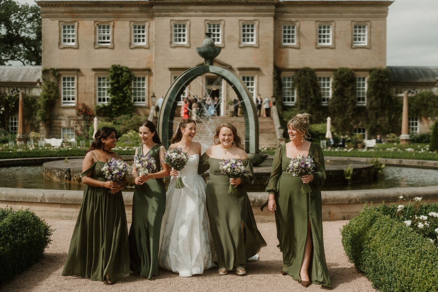 Bride and bridesmaids in sage green gowns holding bouquets in a formal garden with a grand manor house backdrop.