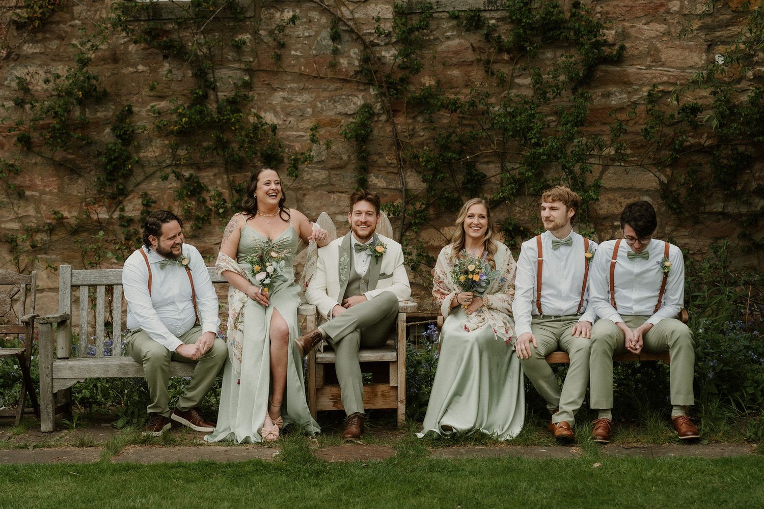 Wedding party laughing on a bench against a stone wall, bride in white, bridesmaids in sage green, groomsmen in suspenders.