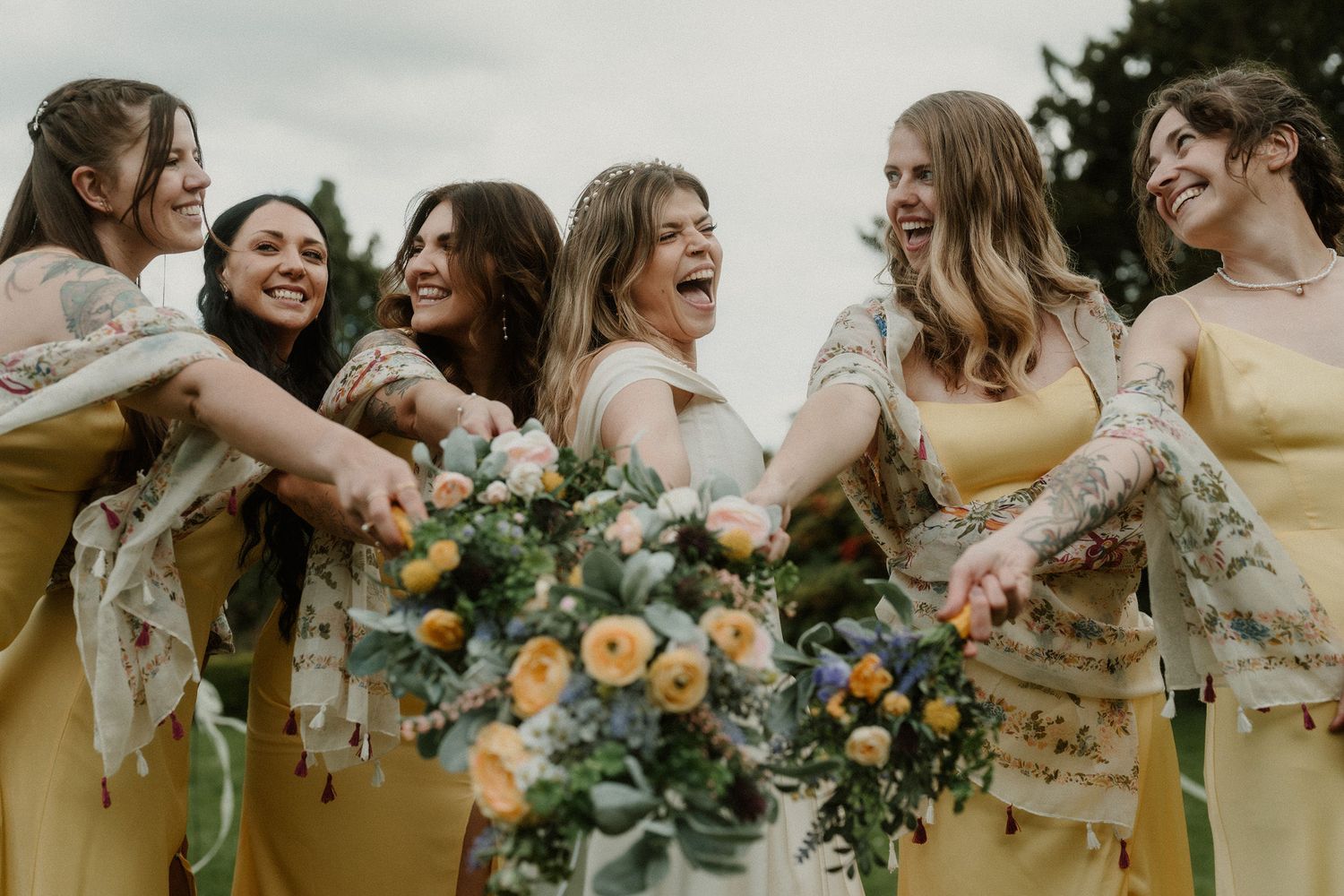 Six bridesmaids in yellow dresses laughing and holding colorful bouquets at an outdoor wedding celebration.