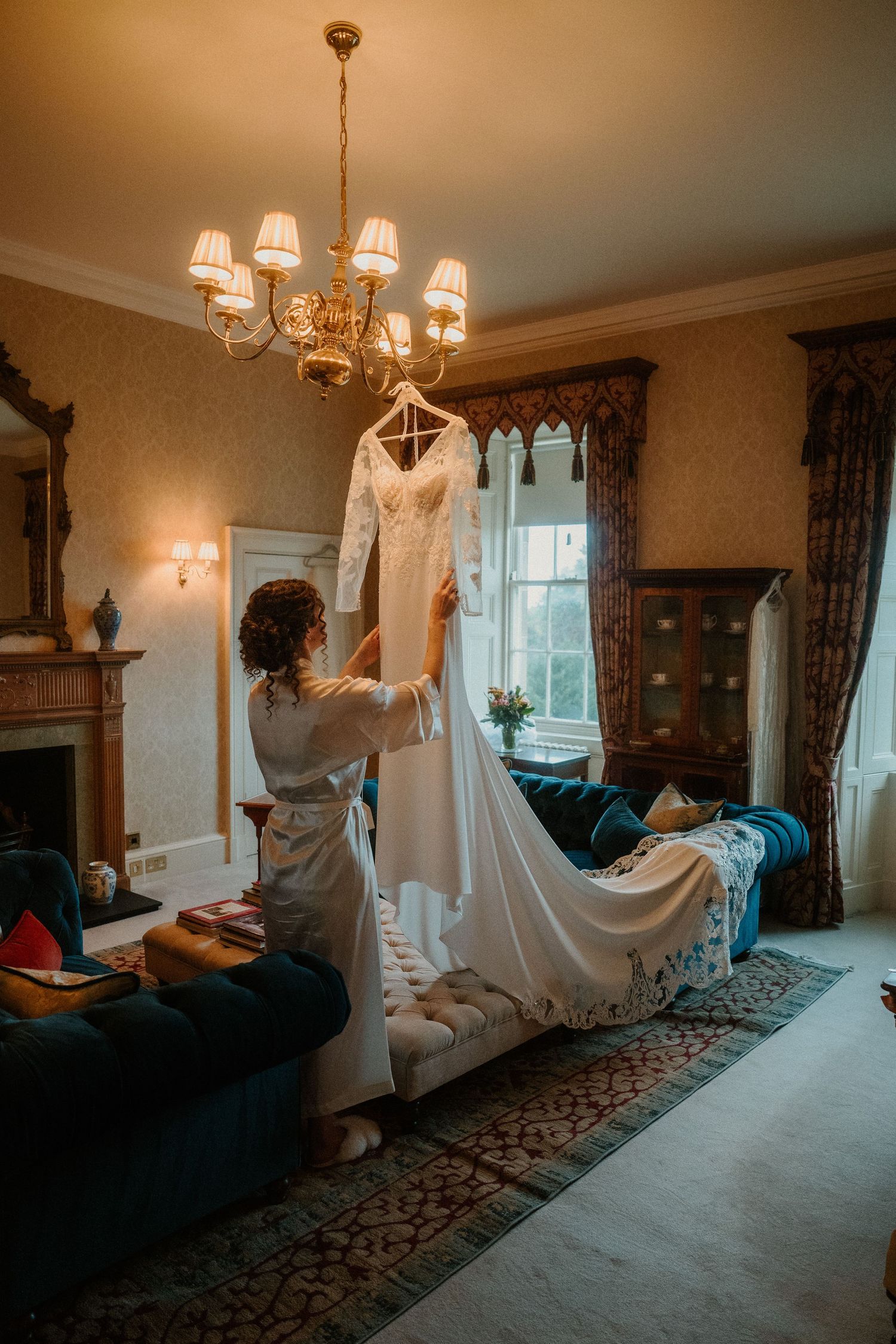 Bride in robe holding white lace wedding dress on hanger in elegant room with chandelier and fireplace.