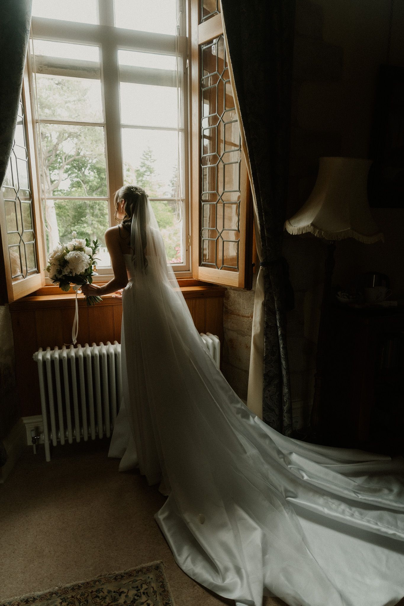 Bride in white gown and veil stands by ornate window holding bouquet, cathedral train flowing on floor.