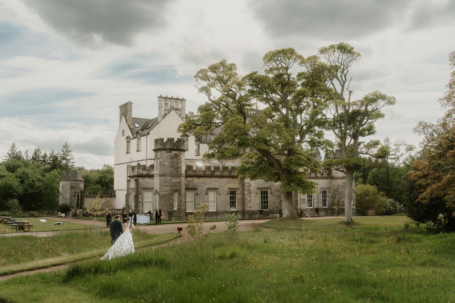 Wedding couple poses before a grand Scottish castle venue surrounded by lush green grounds and tall trees.