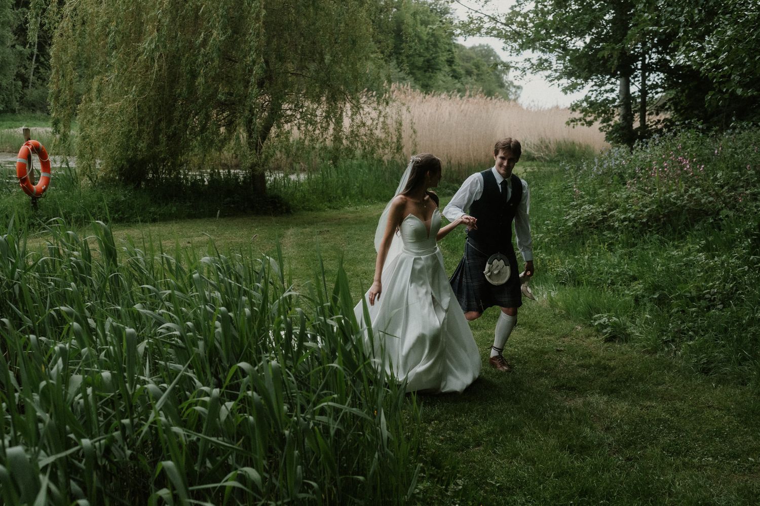 Bride and groom walking through lush green gardens, groom wearing traditional Scottish kilt, on their wedding day.