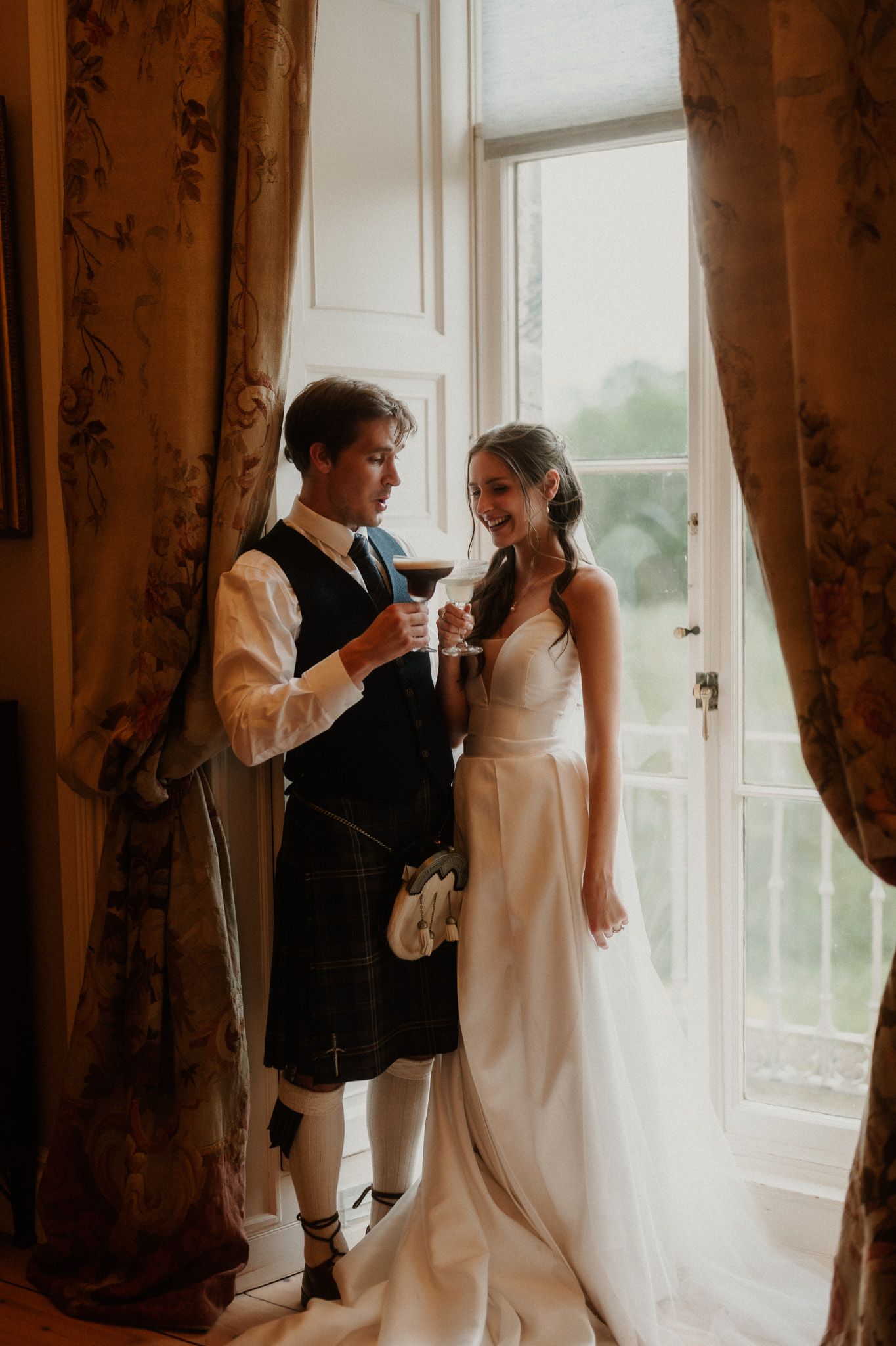 A bride in a white gown and groom in a Scottish kilt share a toast by a sunlit window with floral curtains.