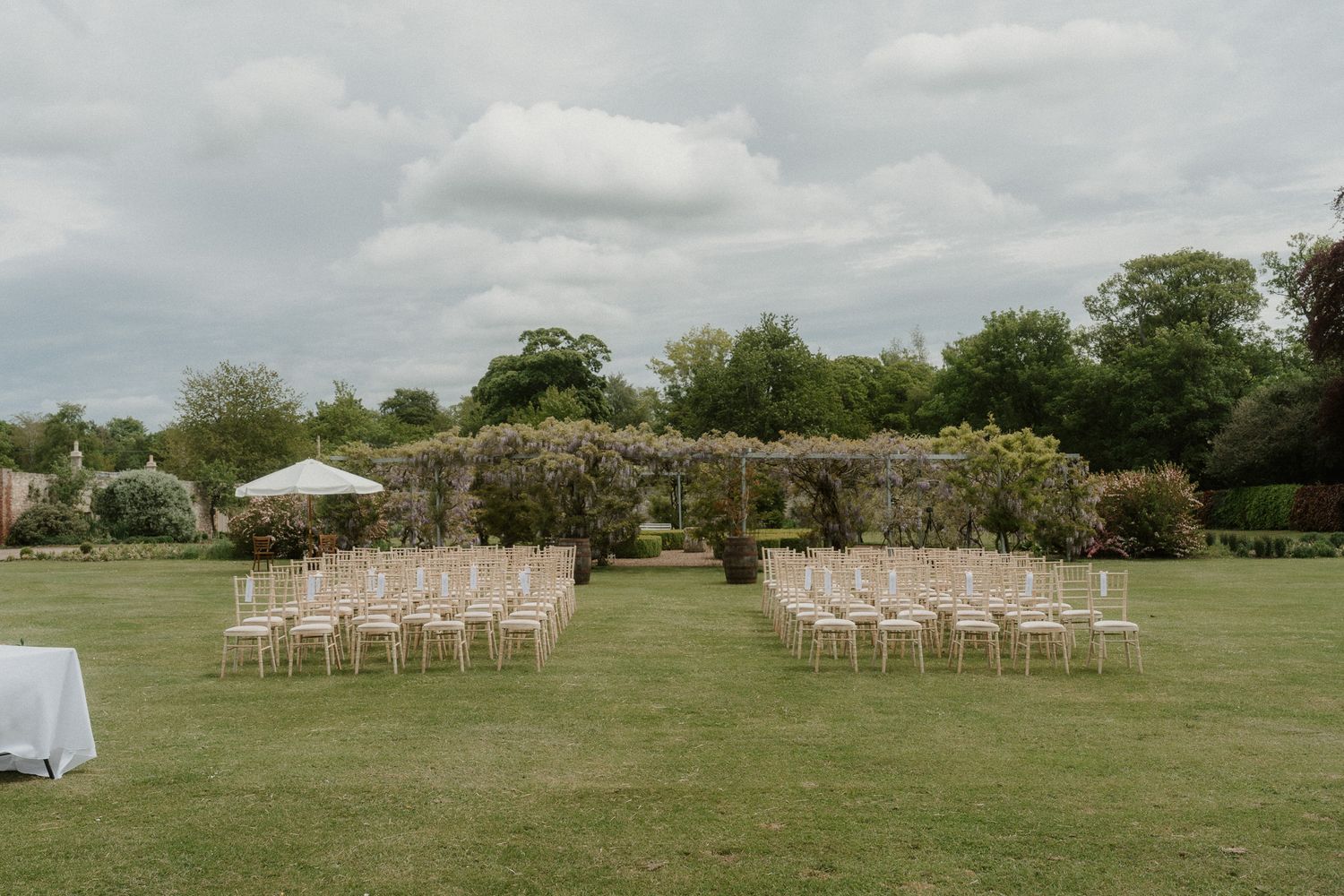 Outdoor wedding ceremony setup with elegant chairs on lush green lawn, floral arch backdrop, and cloudy sky.