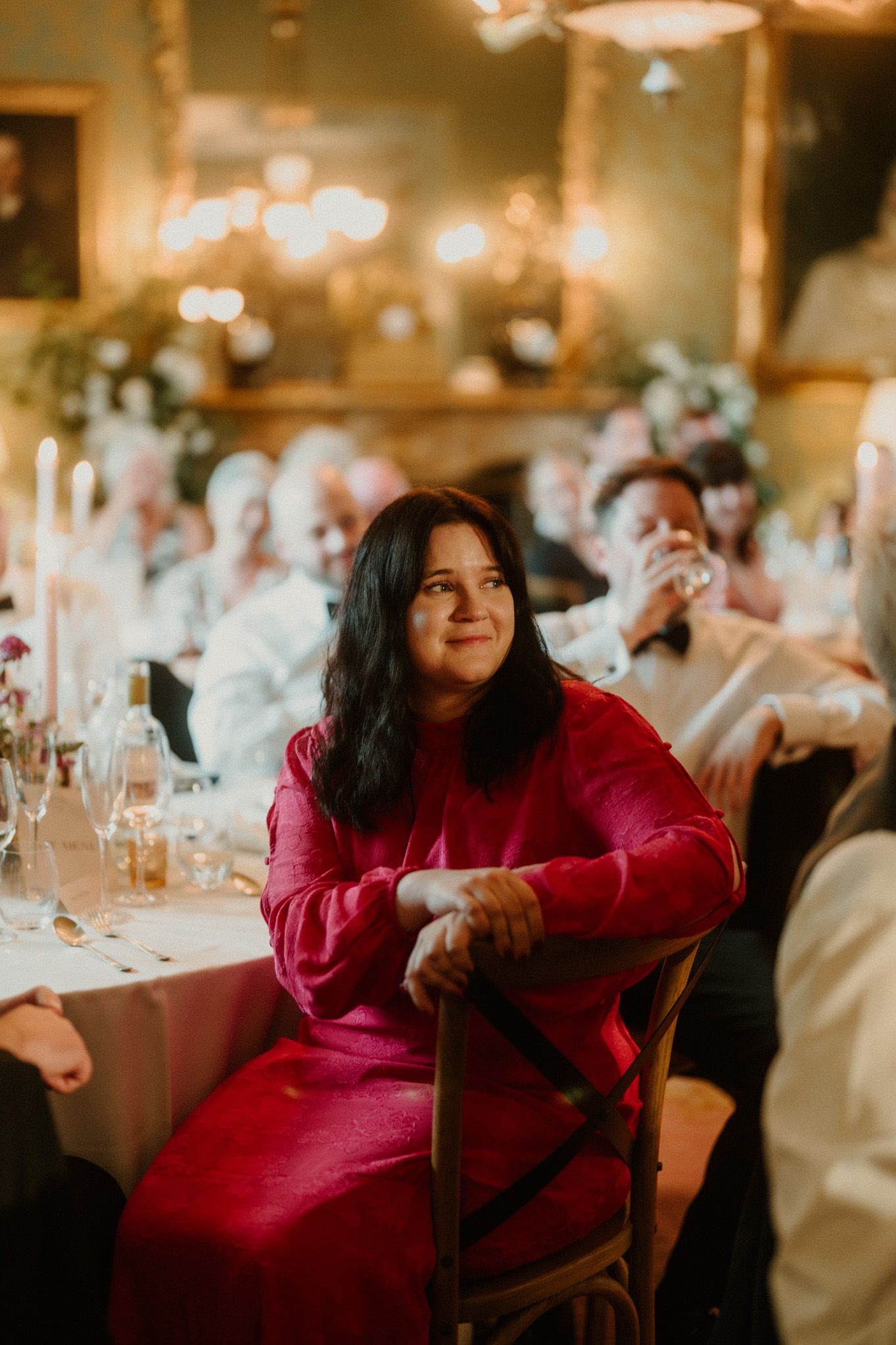 A smiling woman in a red dress sits on a wooden chair at an elegant candlelit wedding reception as guests enjoy the speeches.