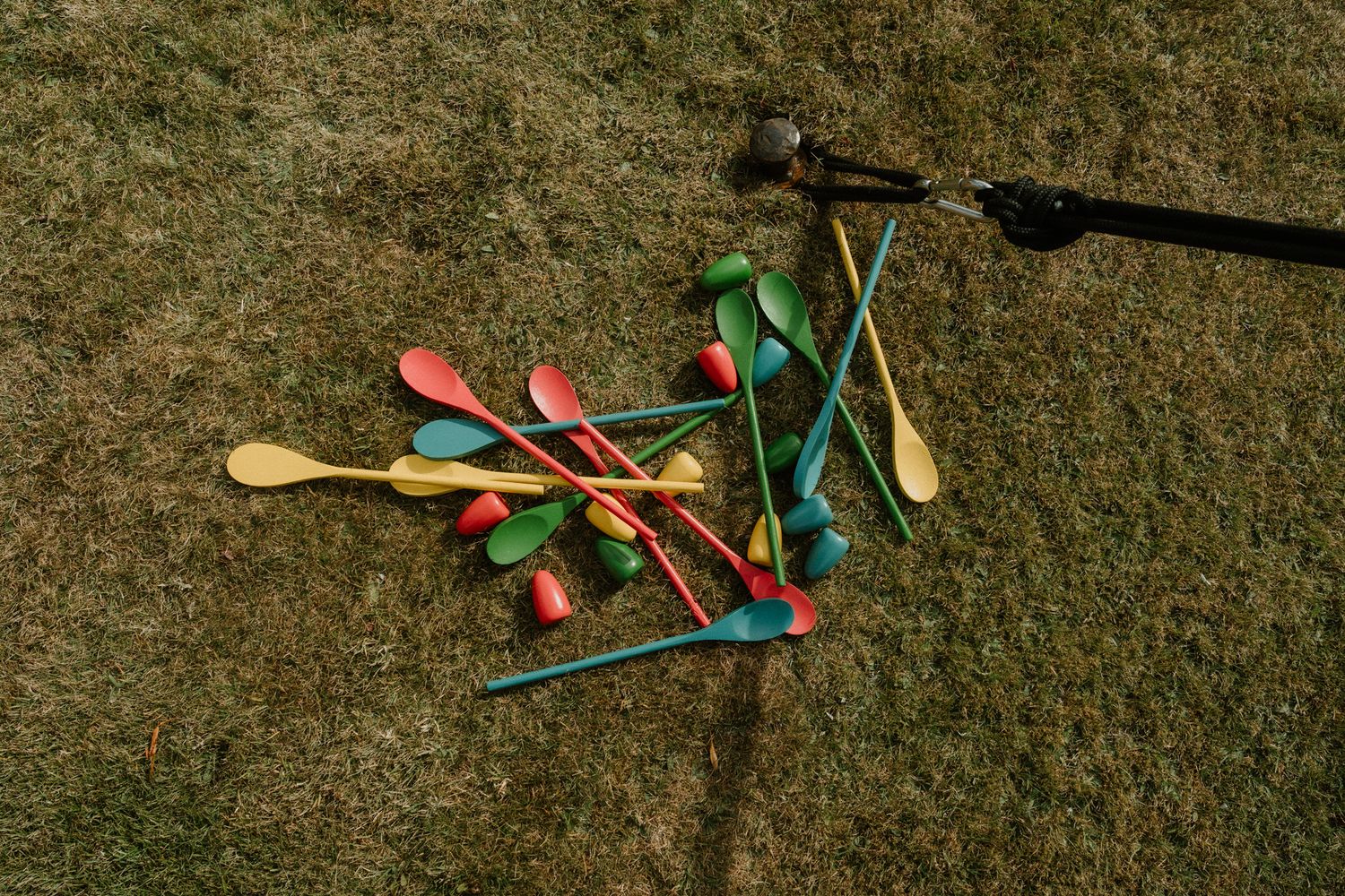 Colorful plastic spoons in red, green, blue and yellow scattered across dry grass with a shadow of a person holding a camera nearby.