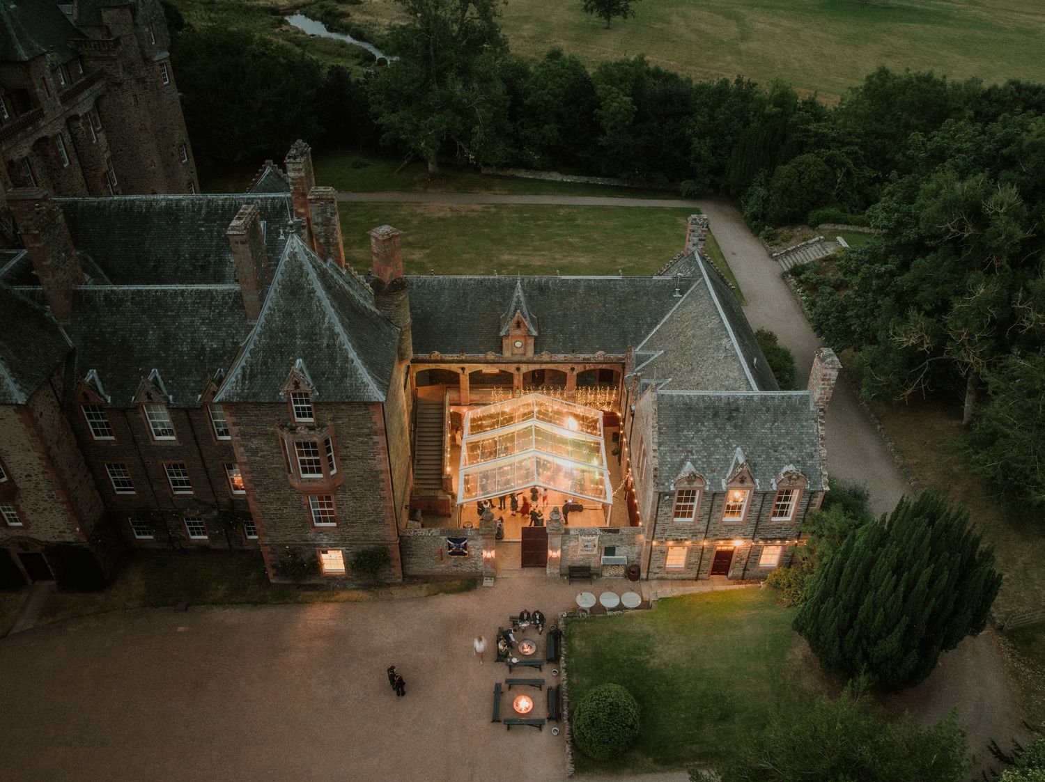 Aerial view at dusk of a historic stone castle with a glowing illuminated courtyard marquee, ideal for a luxury wedding venue.