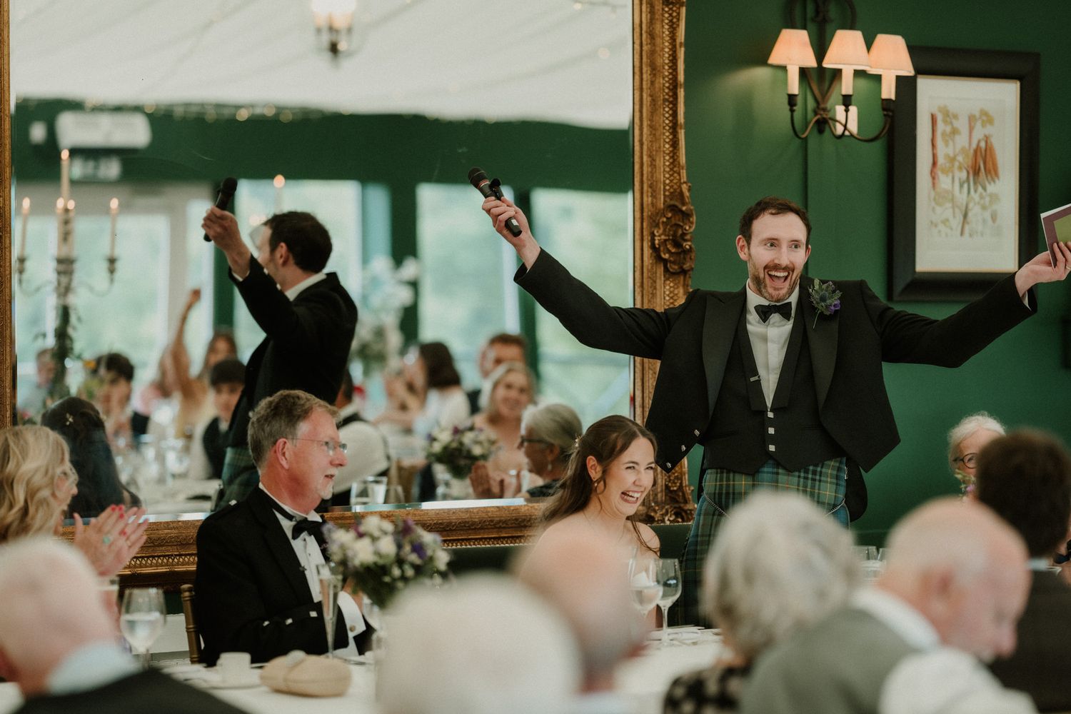 Joyful wedding reception toast with groom in kilt raising arms, guests celebrating at elegantly decorated tables in green venue.