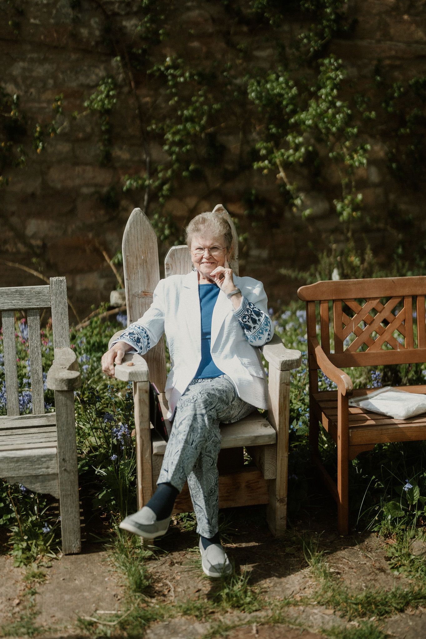 Elderly woman in white blazer and blue pants sitting in wooden garden chair, smiling outdoors.