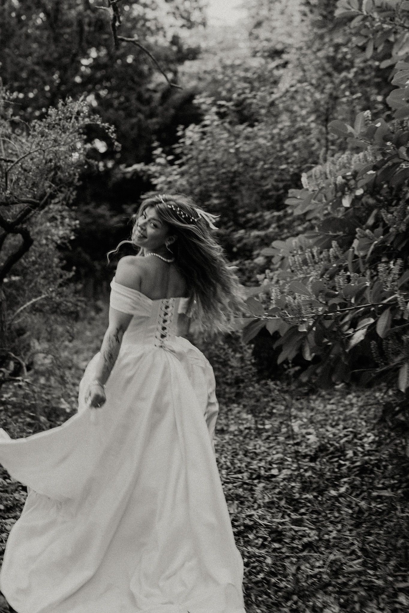 Bride in white off-shoulder ball gown with floral crown twirling in black and white forest setting.