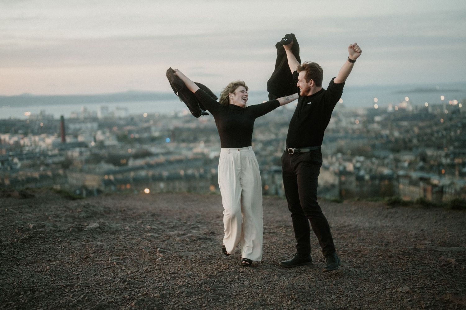 Couple dancing joyfully on hilltop at dusk with city skyline backdrop, arms raised holding clothing.