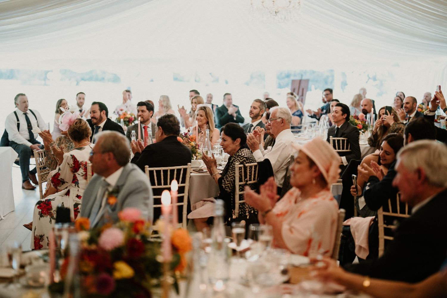 Wedding guests celebrate during reception toasts in a decorated white tent venue.