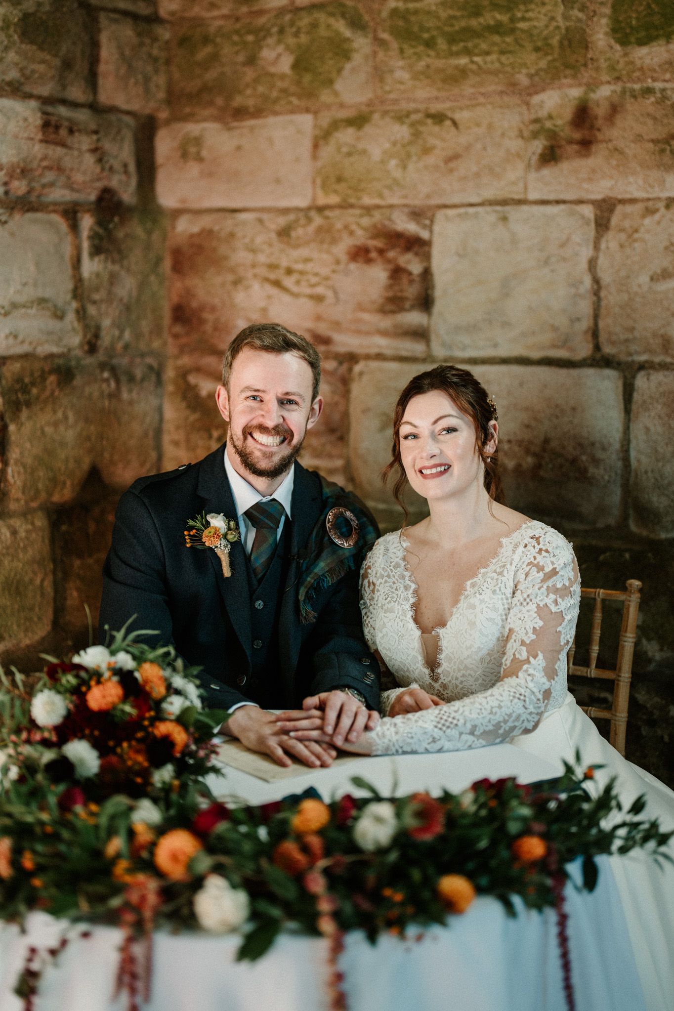 Newlyweds seated at table with autumn-colored floral arrangements in stone castle setting.